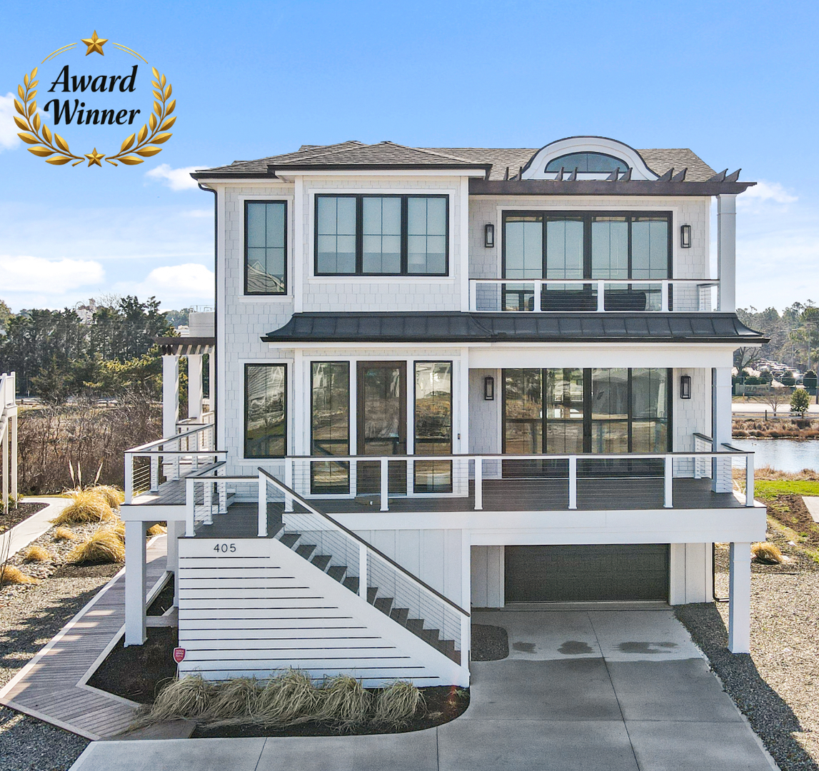 Modern two-story white beach house with large windows, balcony, and driveway overlooking water
