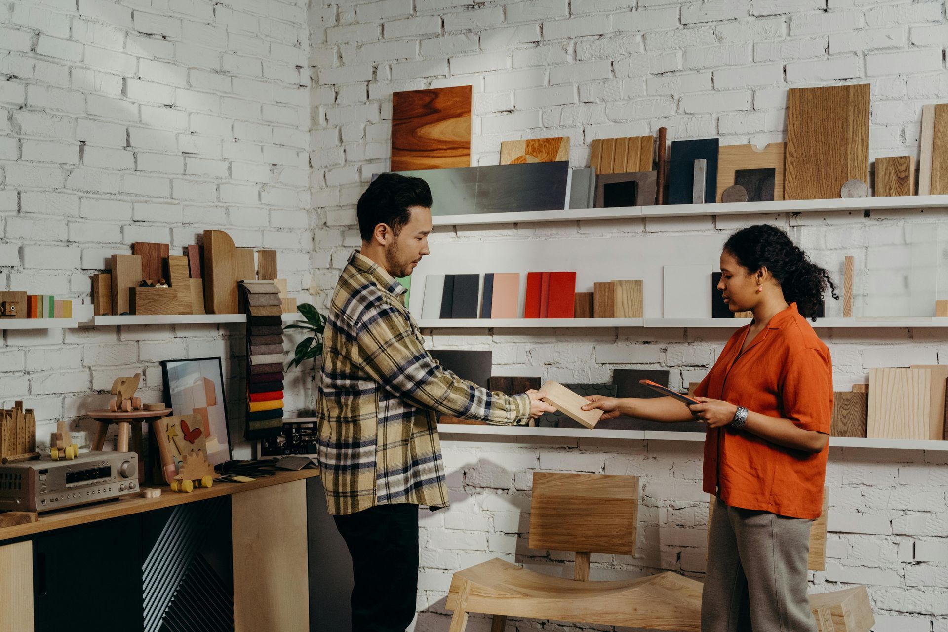 A man and woman shake hands in a wood shop, shelves filled with wood samples.