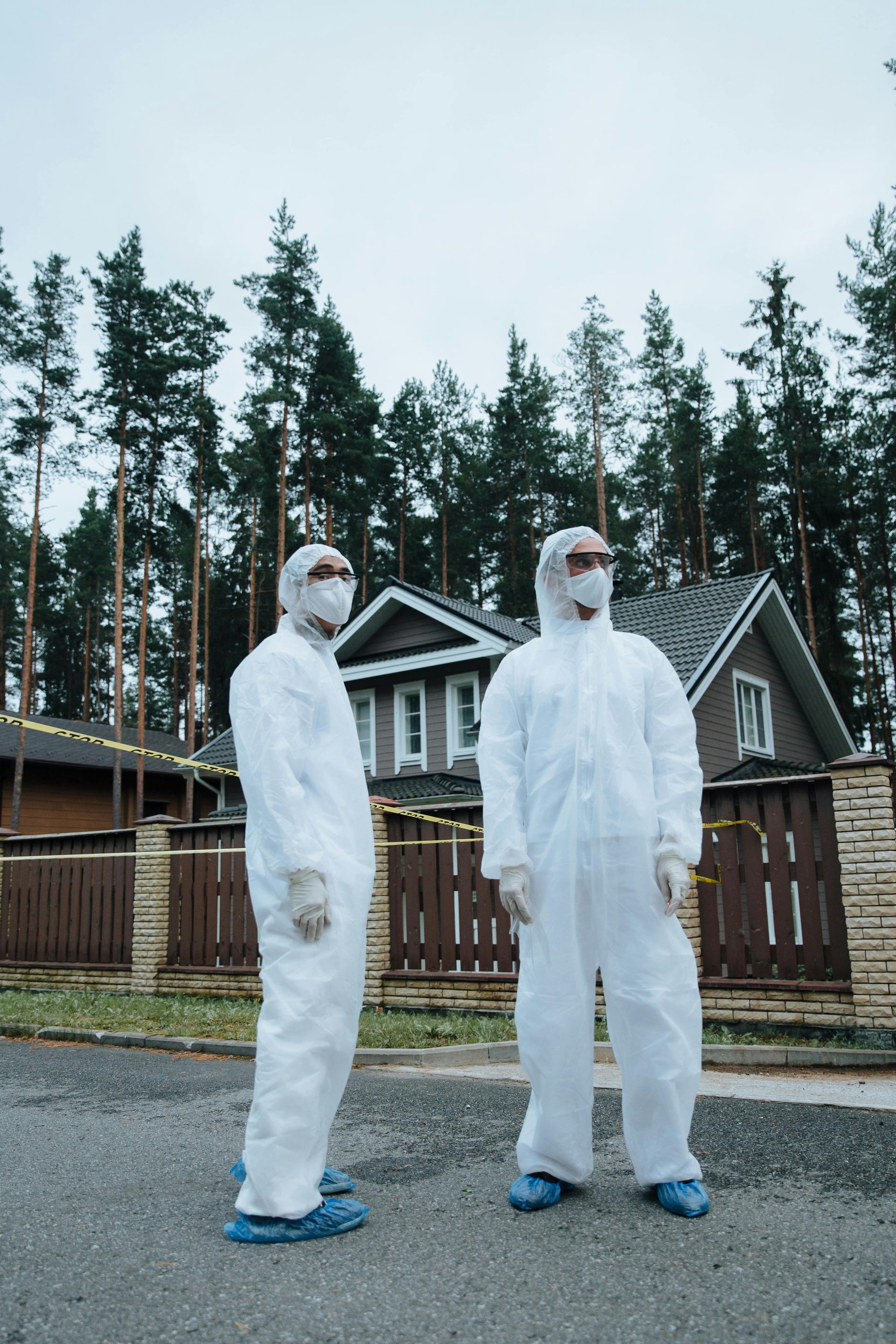 Two people in white protective suits and blue shoe covers stand on a gravel driveway in front of houses and trees.