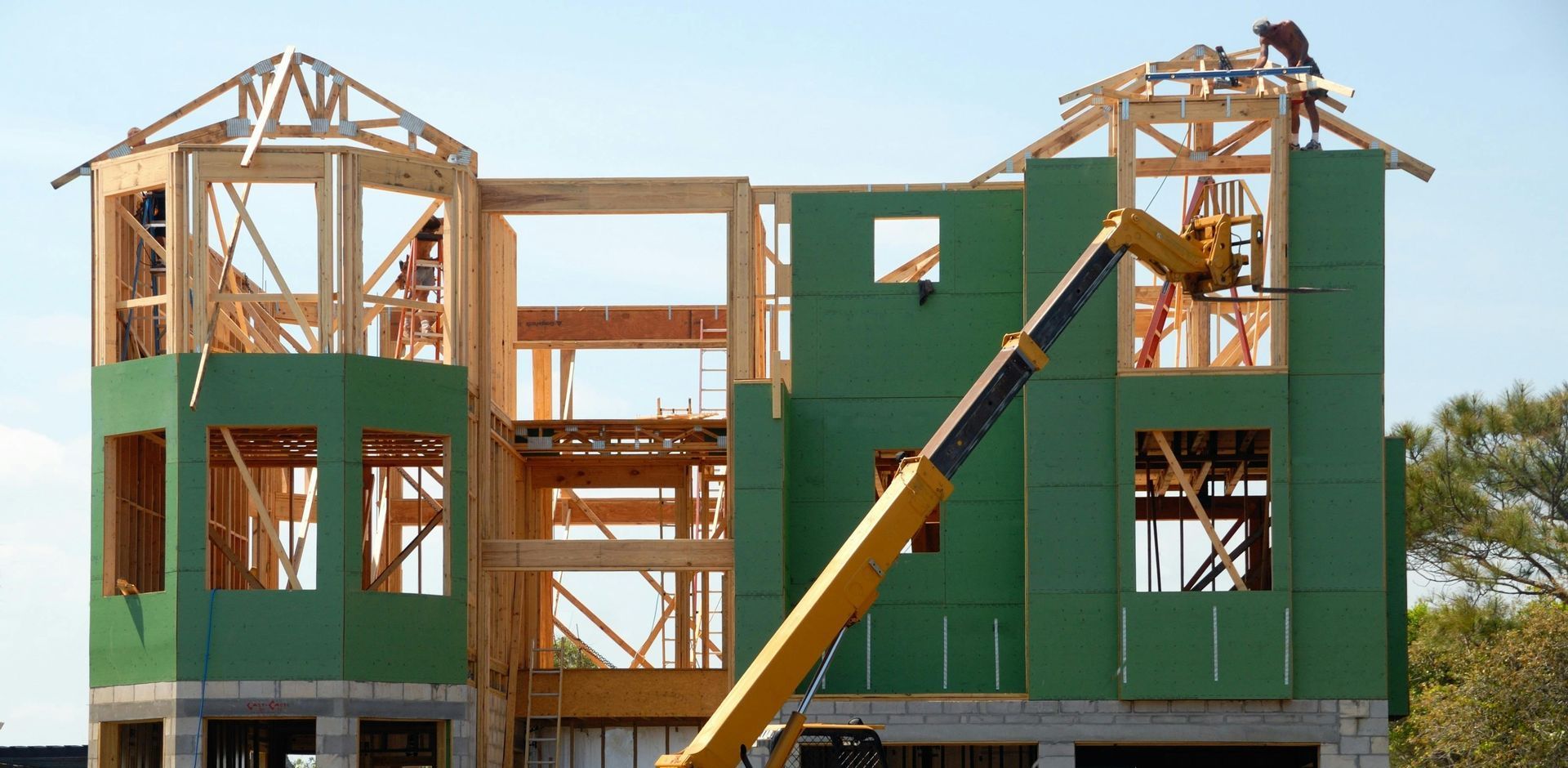Construction site of a multi-story house with green siding and a yellow lift; two workers are visible.