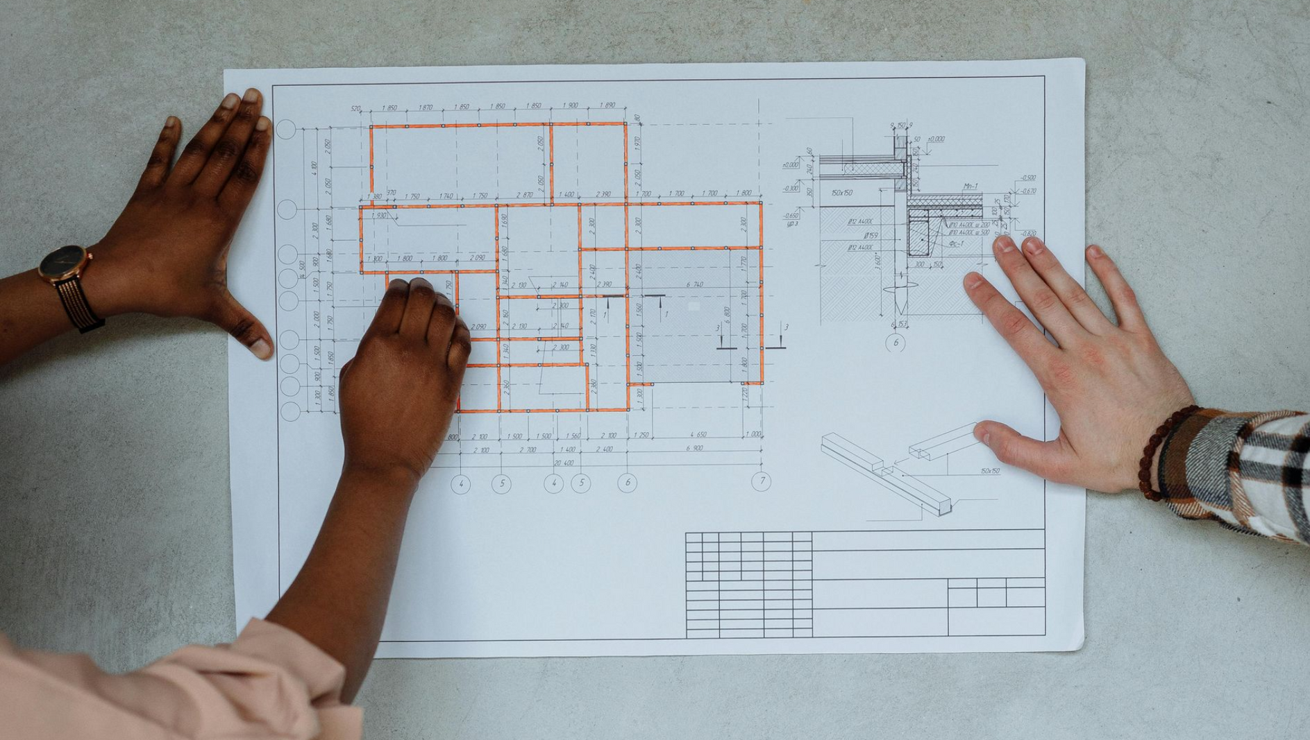 Two hands reviewing an architectural blueprint with orange markings on a white table.