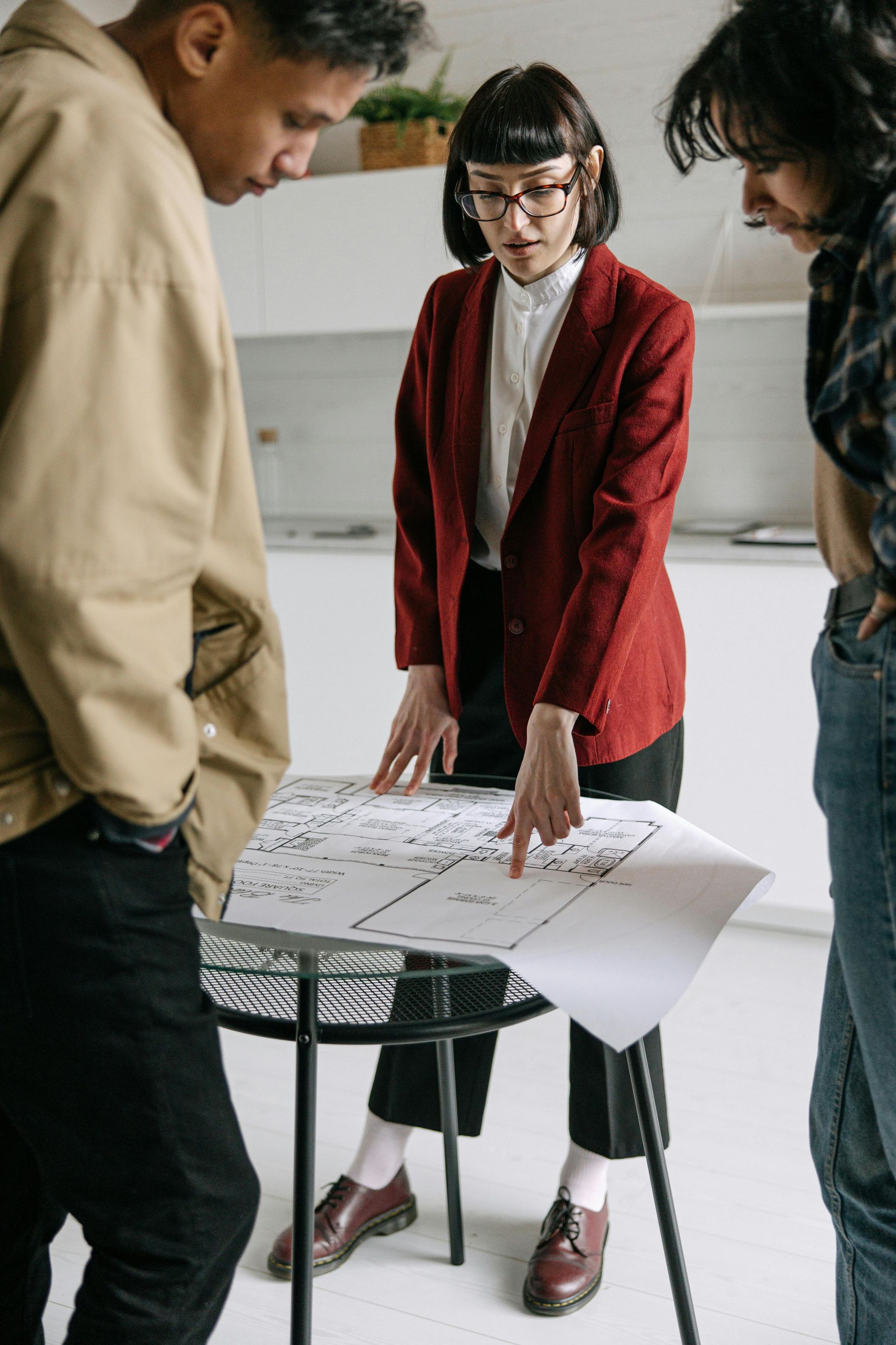 Three people around a table, looking at a blueprint. Woman in red blazer points. Light-filled room.