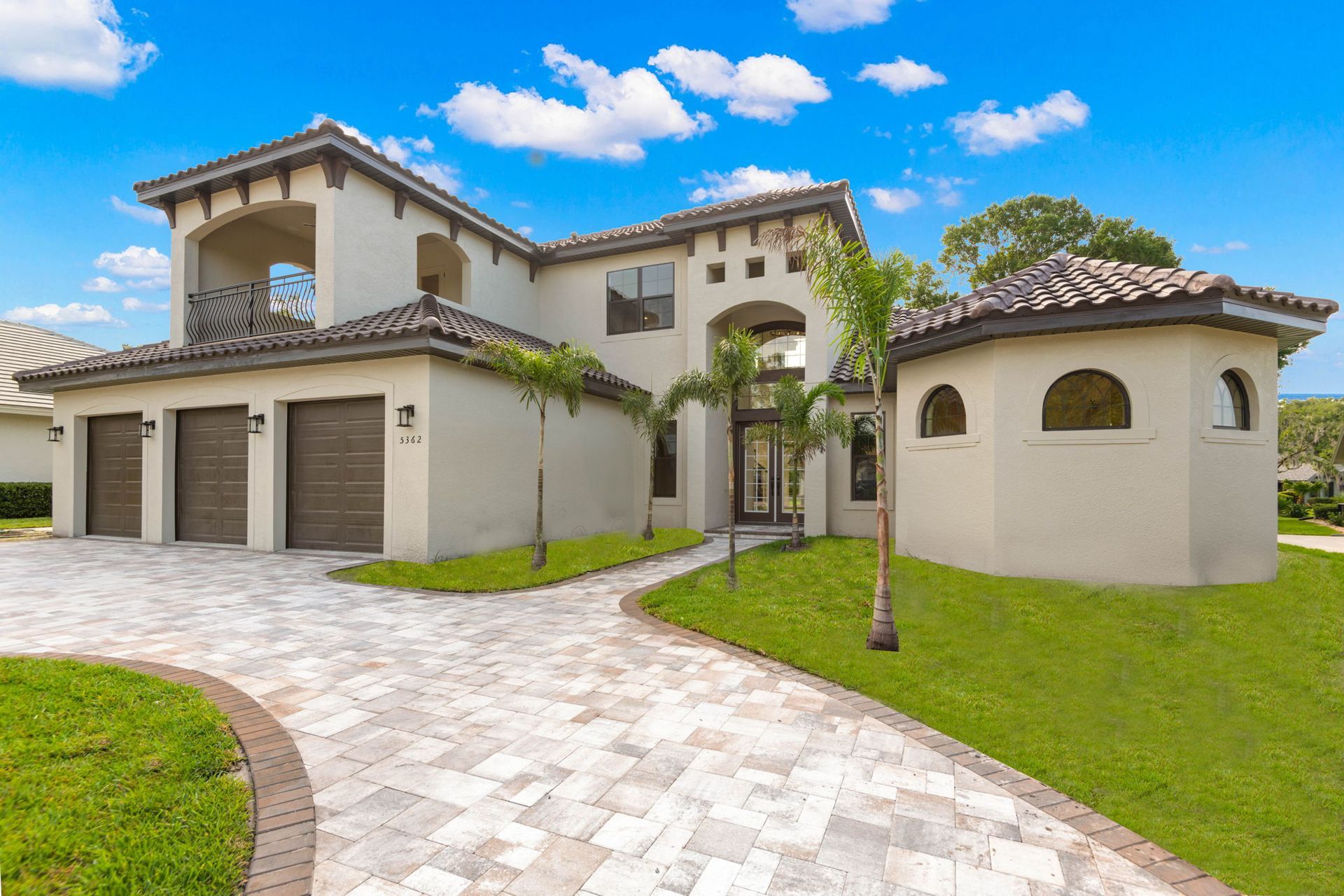 Luxurious beige two-story home with tile roof, arched entryway, palm trees, and brick driveway under blue sky.