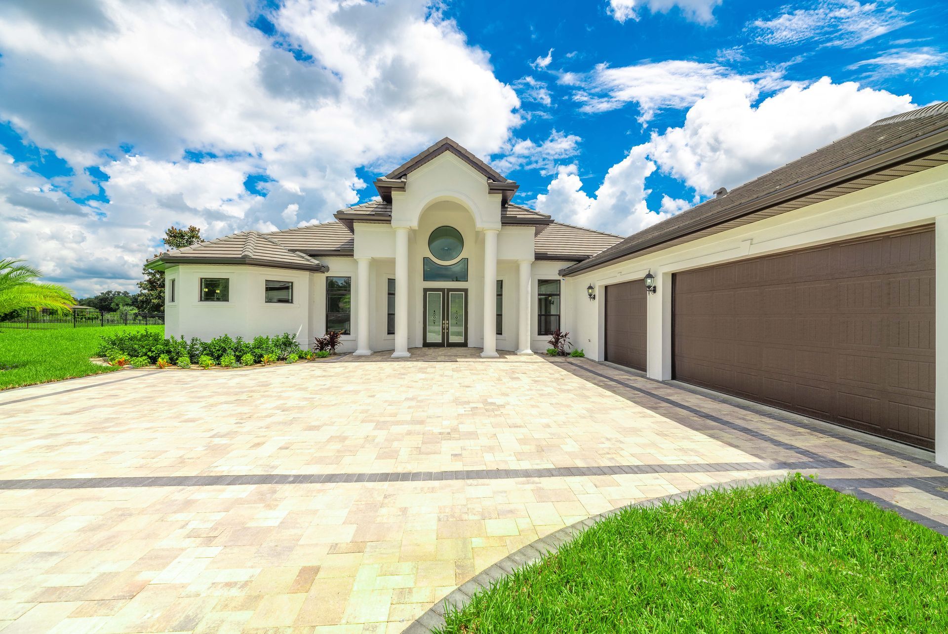 Large white house with columns, brown garage doors, and paver driveway. Sunny day.
