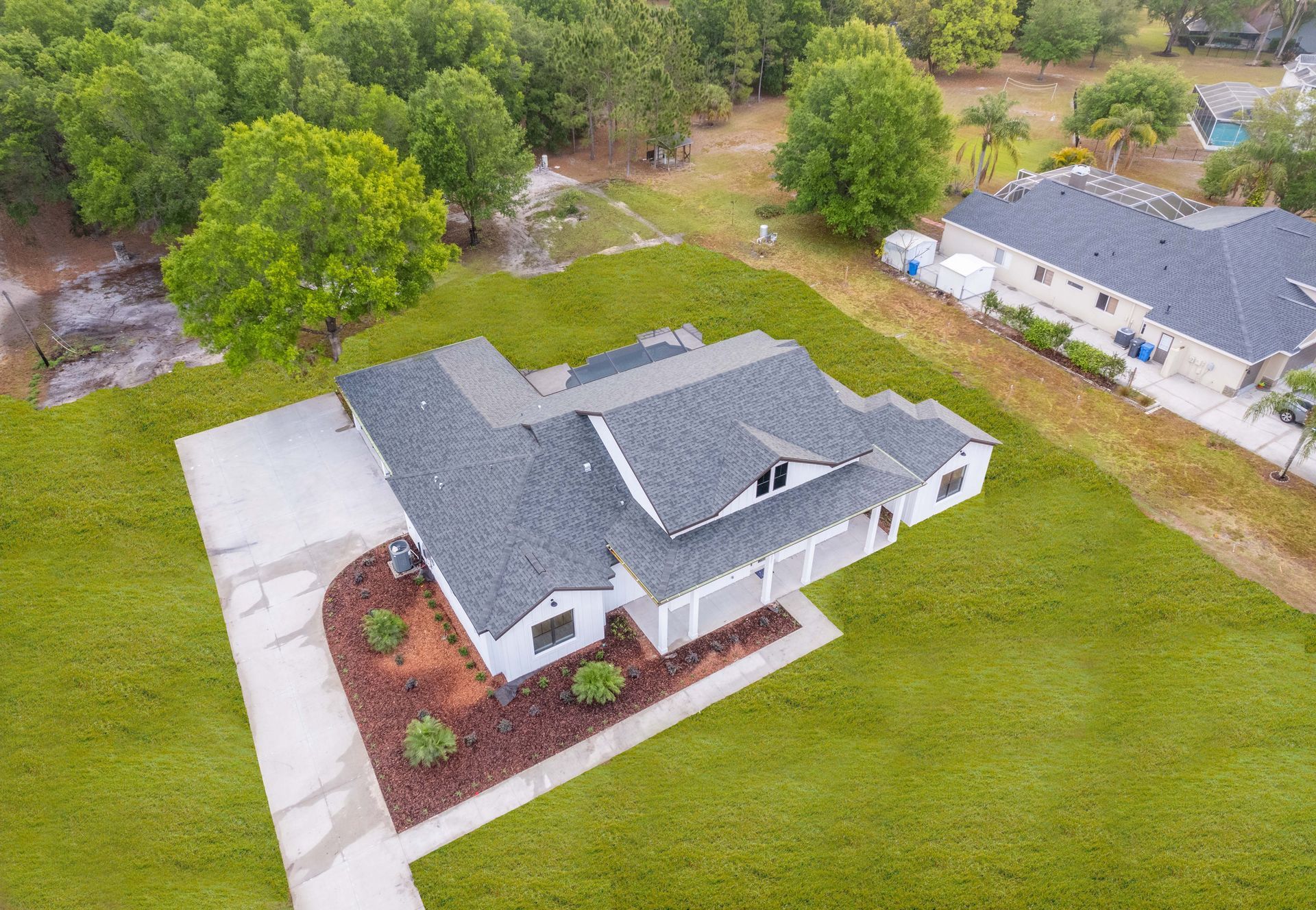 Aerial view of a gray-roofed house with a concrete driveway on a green lawn, adjacent to trees and another building.