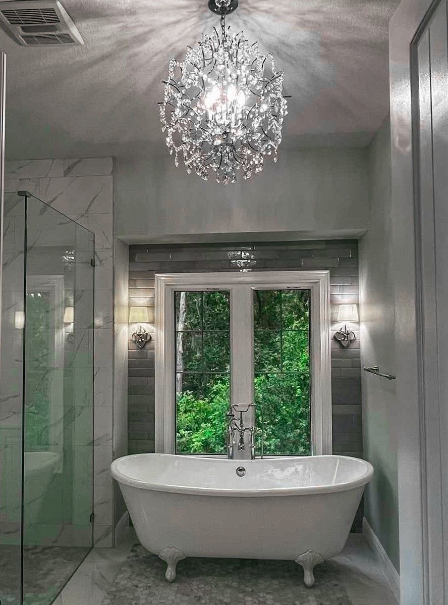 Elegant bathroom with a clawfoot tub, chandelier, and a window overlooking greenery.