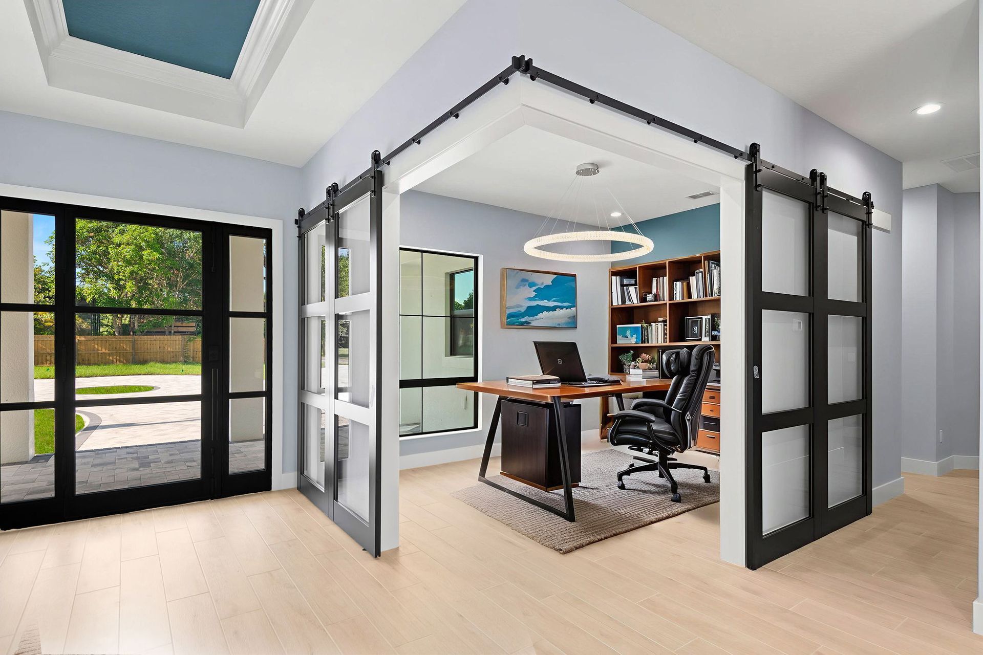 Office with barn-style doors; light wood floors, blue-green walls, black framed doors, desk, and a large window.