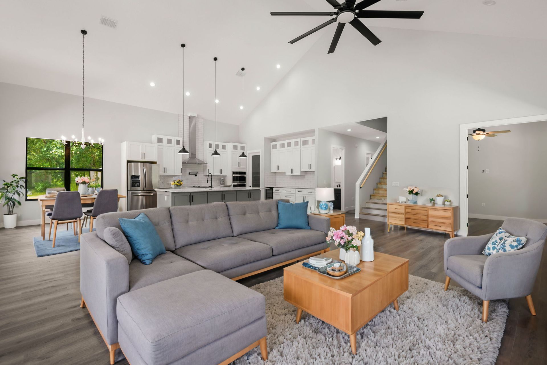 Open-concept living space with a gray sectional sofa, wooden coffee table, and a kitchen visible in the background.
