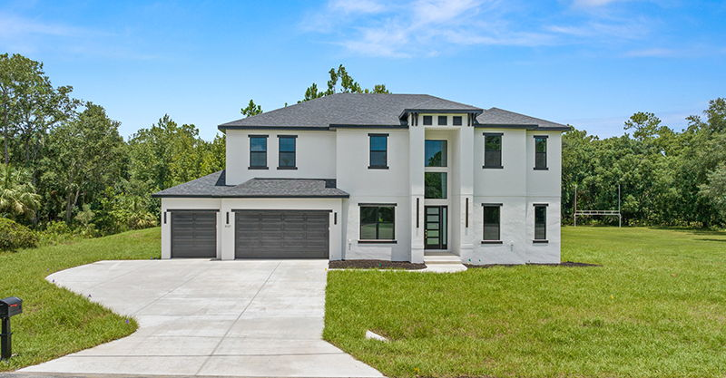 White two-story house with gray roof and garage doors. Driveway leads to the front yard with trees in the background.