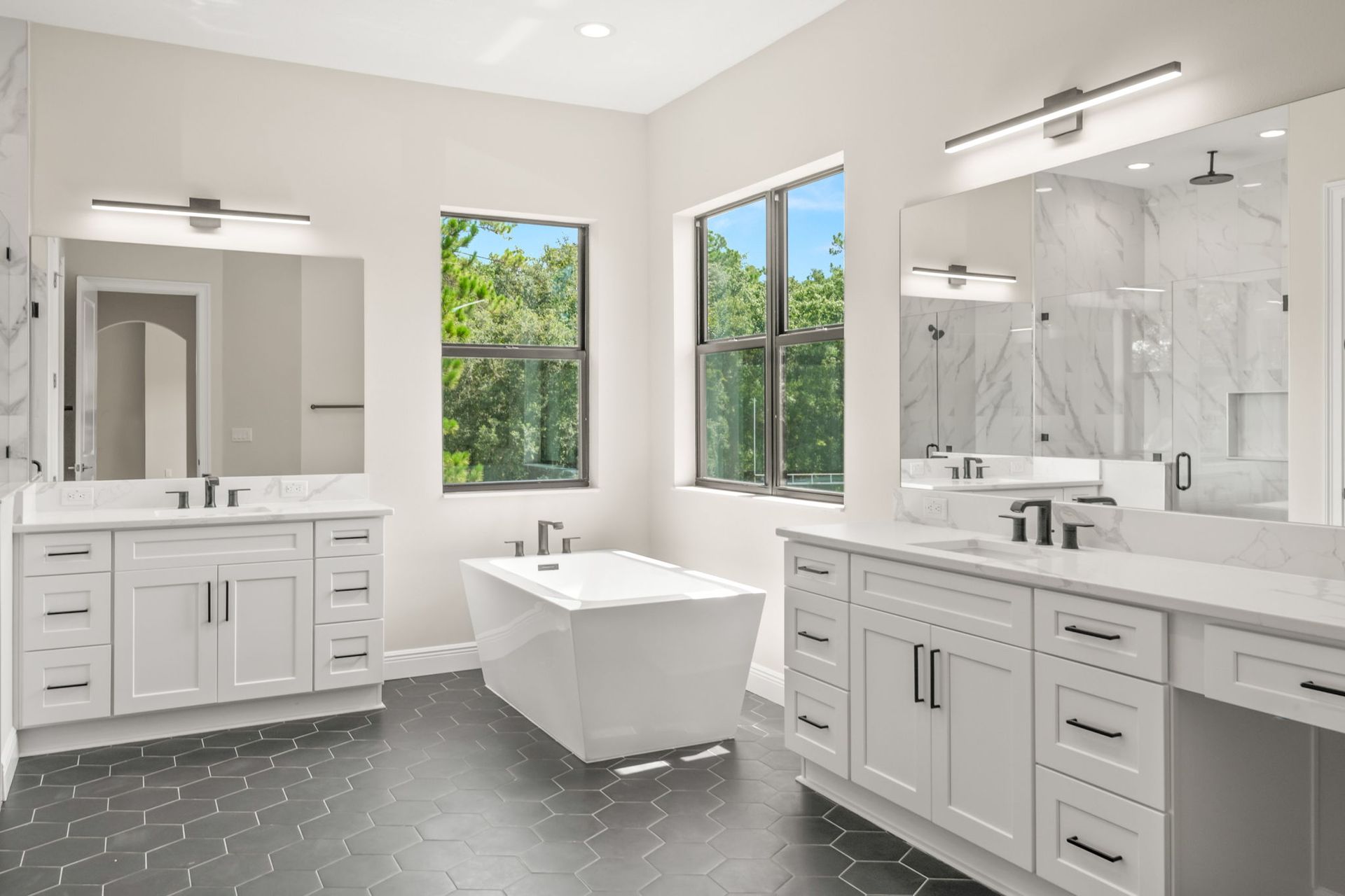 Spacious, white bathroom with double vanity, soaking tub, large windows, and dark hexagonal floor tiles.