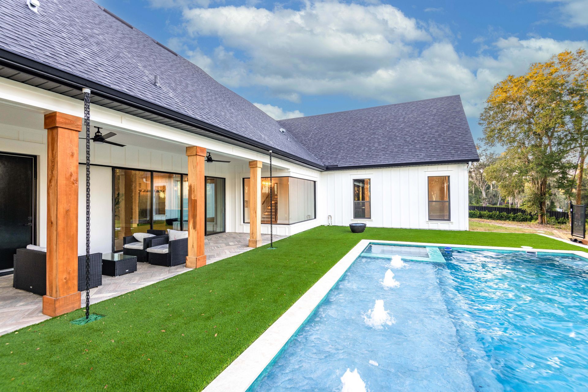 White house with black roof, outdoor pool, patio with furniture, green lawn, blue sky.