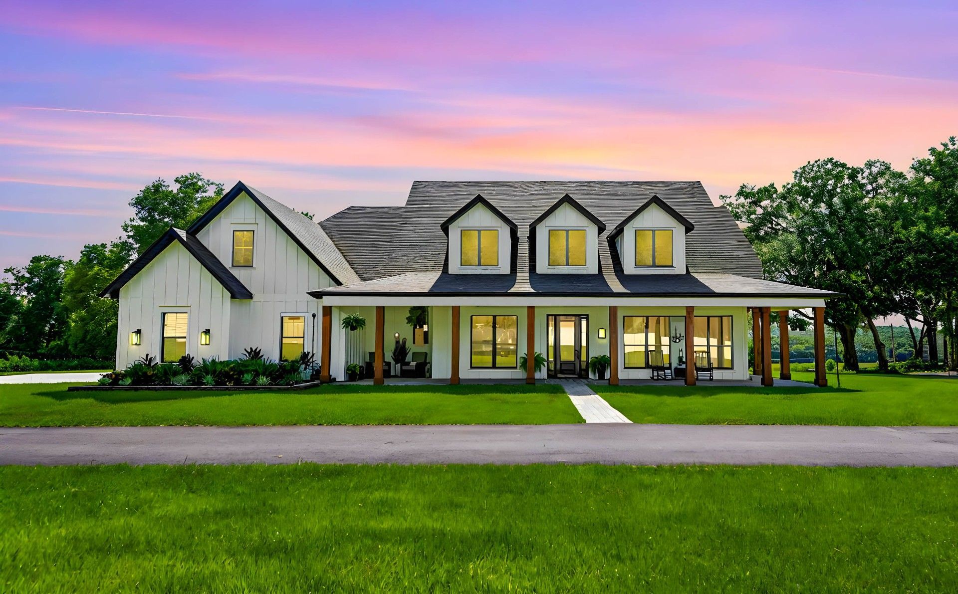 White farmhouse with a long porch, three dormers, and a green lawn under a colorful sunset.