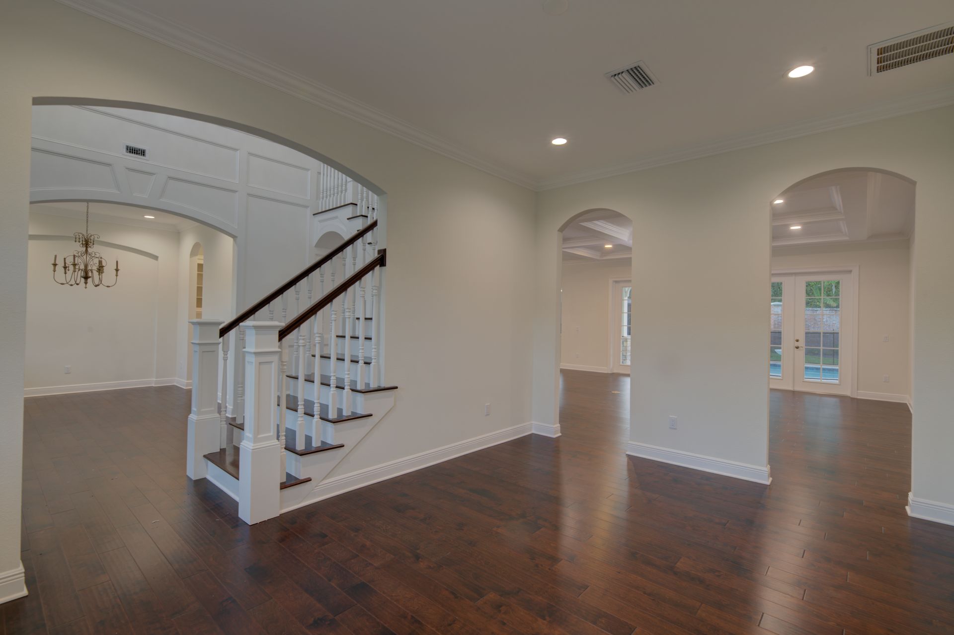 Interior view of a house with dark wood floors, white walls, and arched doorways. Stairs are visible.