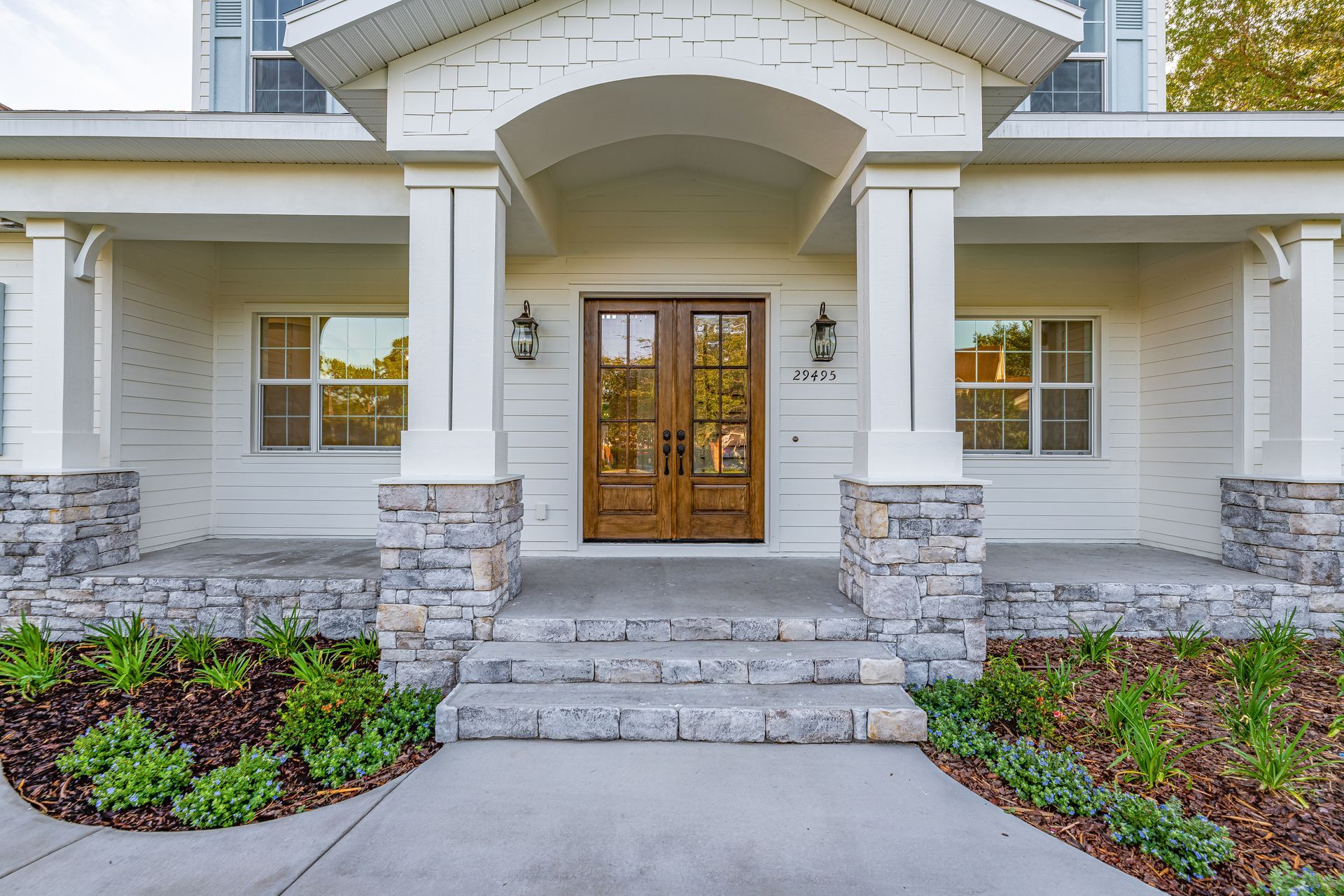 White house with stone accents, arched entryway, double wooden doors, steps, and landscaping.
