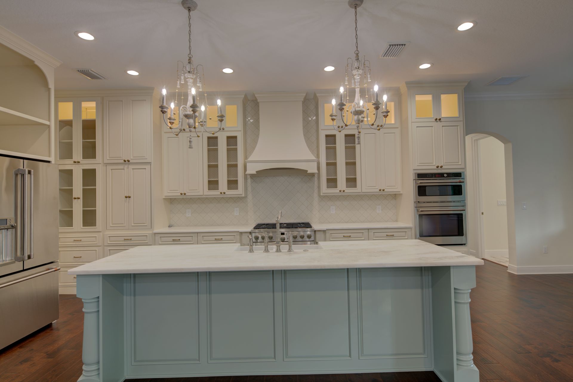 Spacious white kitchen with island, range, and cabinets; dark wood floor, two chandeliers.