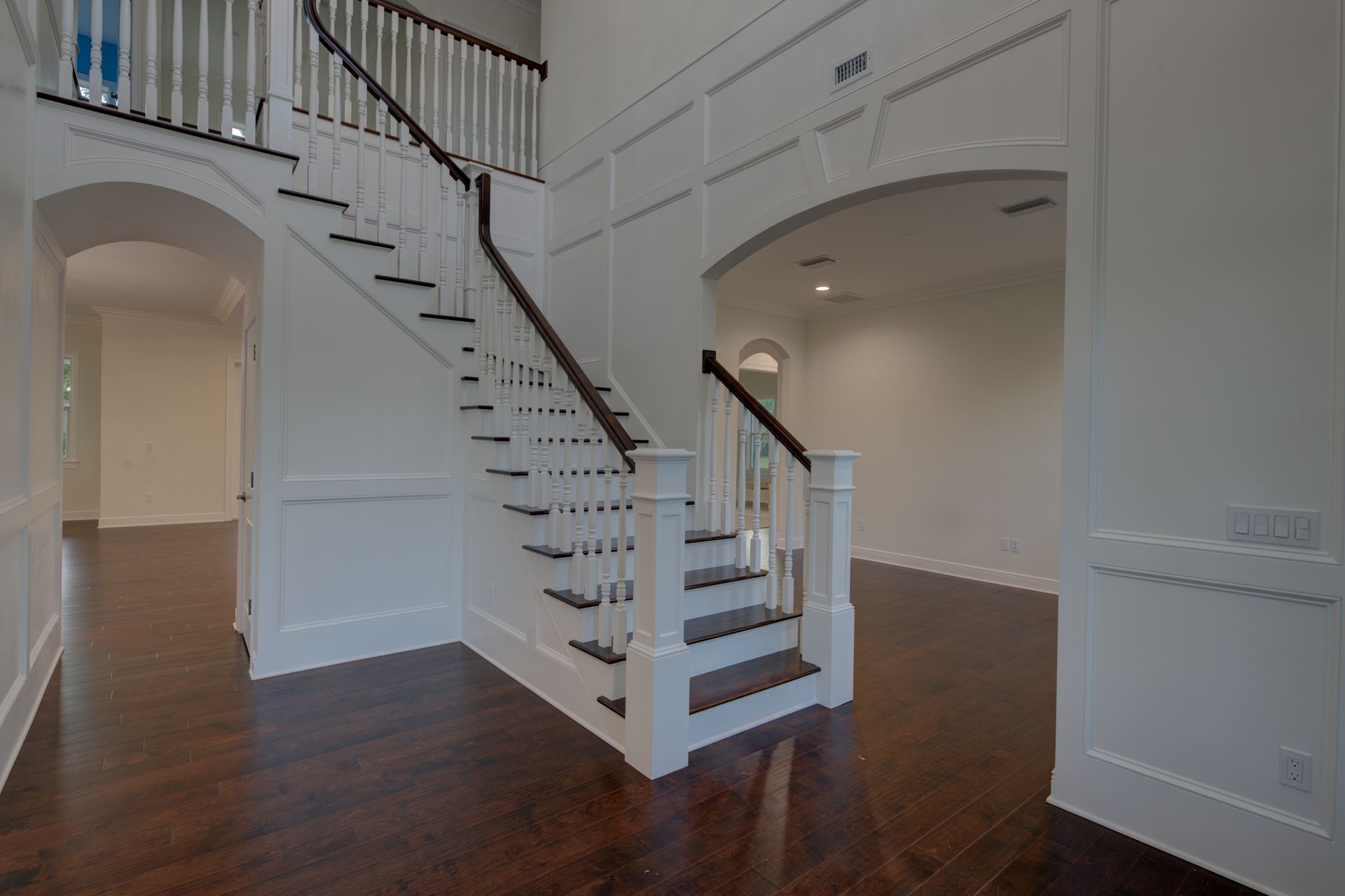 A grand entryway with a staircase, dark wood floors, white walls, and an arched doorway.