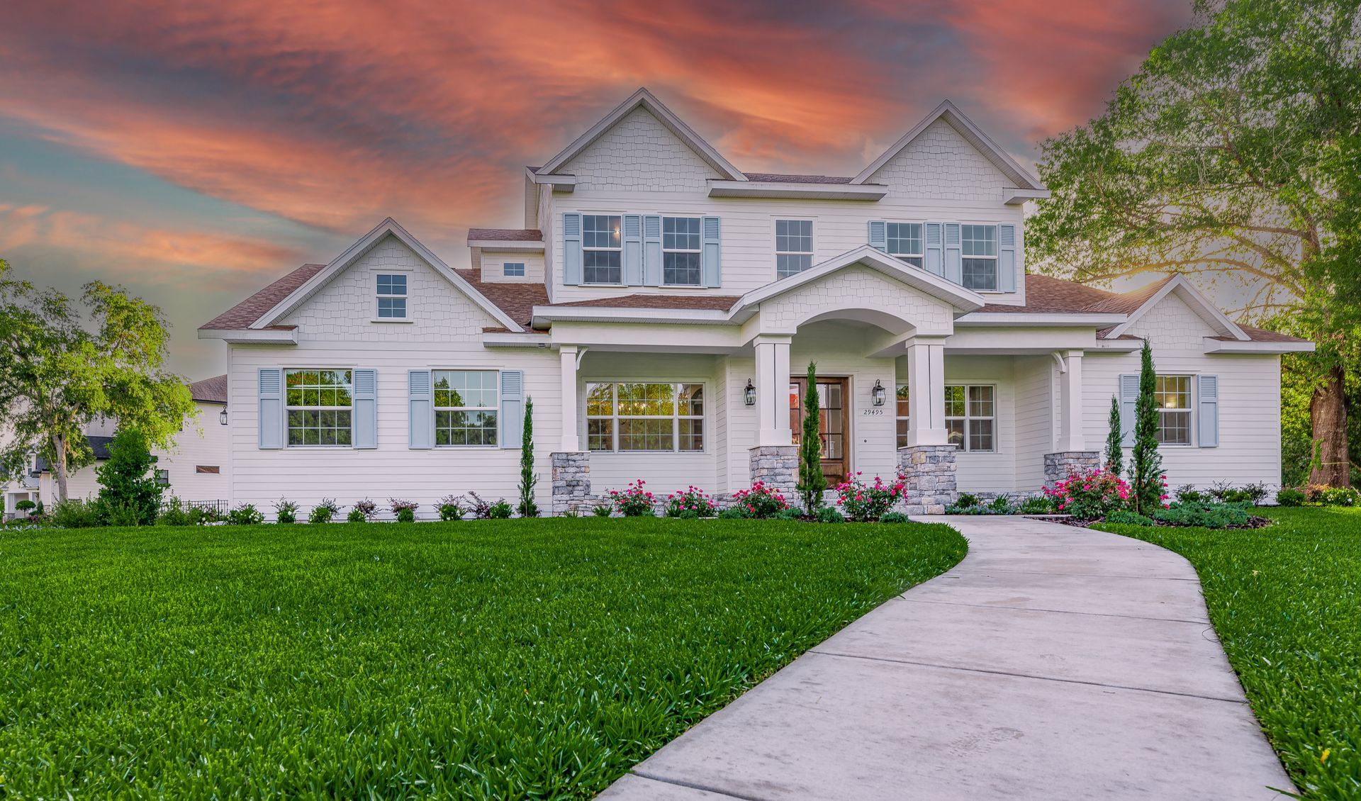 White two-story house with light blue shutters, green lawn, and a winding walkway under a colorful sunset.