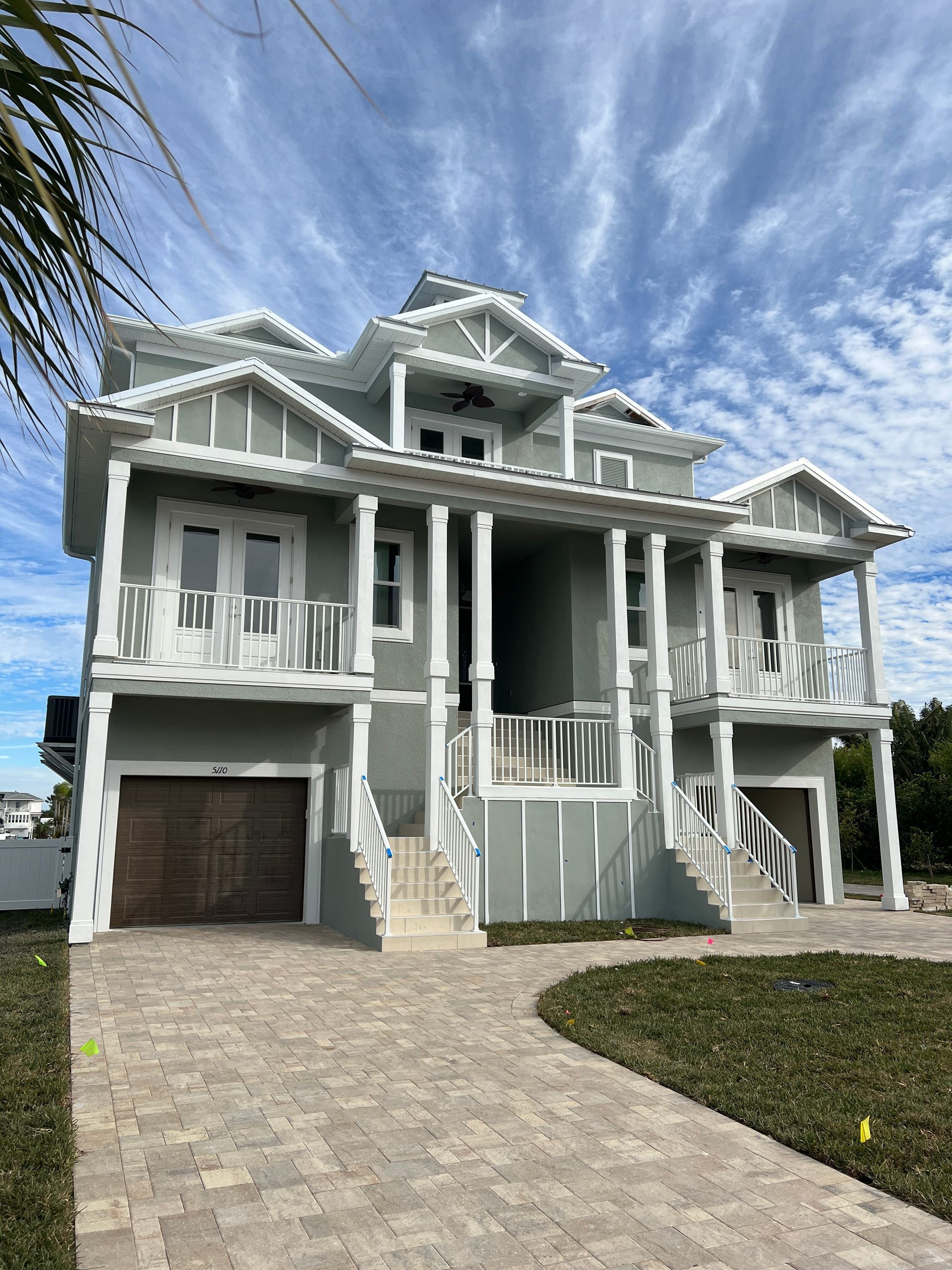 Three-story beach house, light gray siding, white trim, balconies, columns, brick driveway, cloudy blue sky.