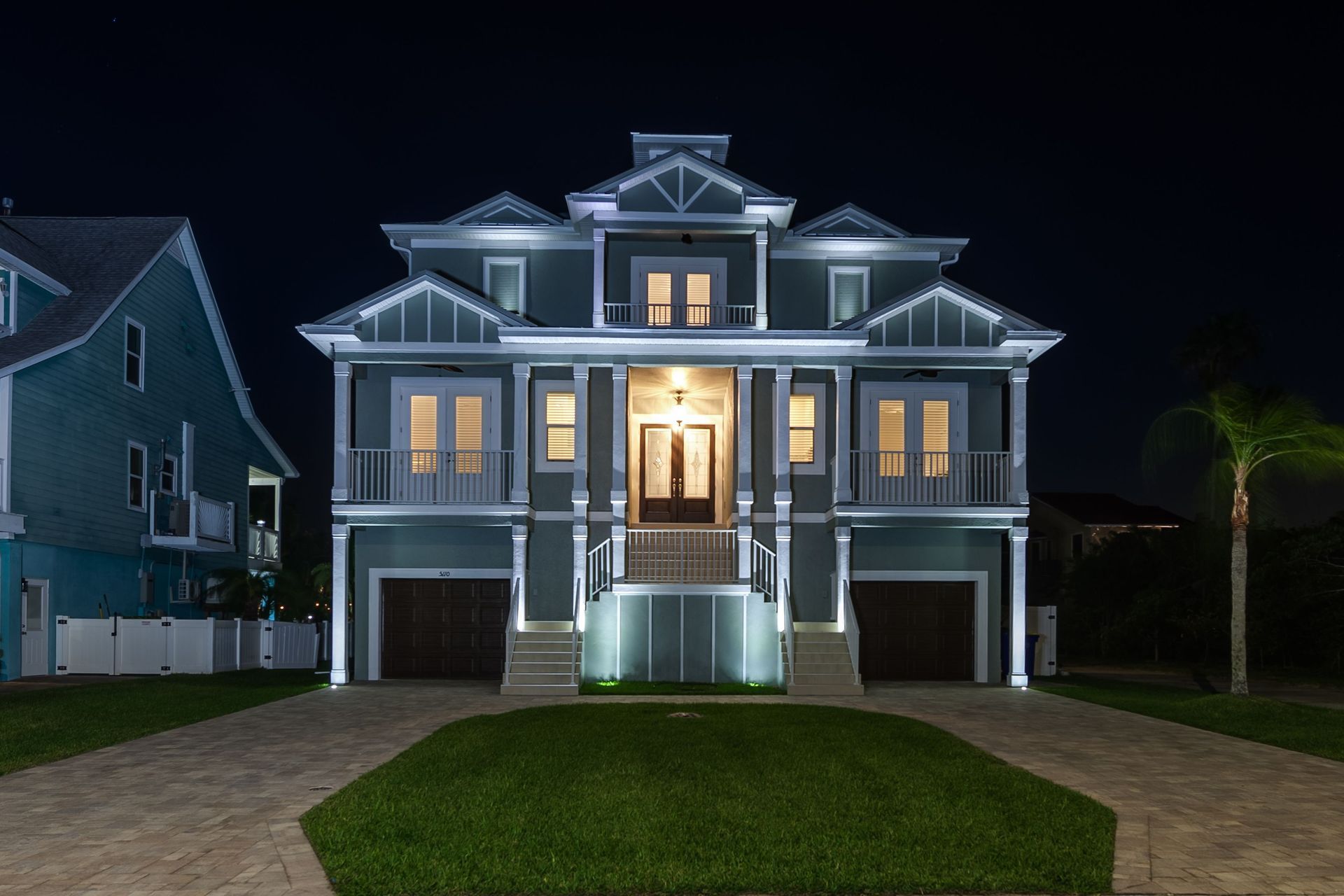 Night view of a multi-story teal house with lit trim and garage doors, fronted by a driveway and grass.