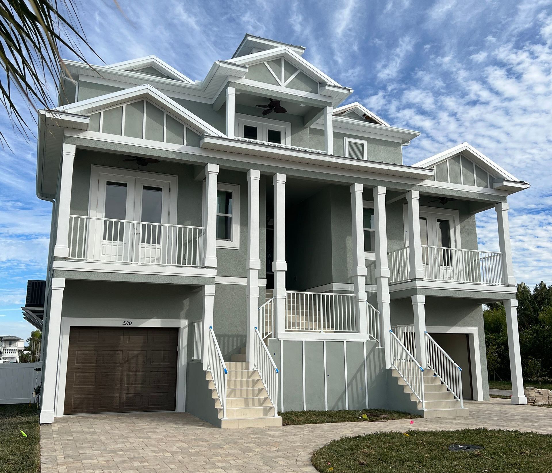 Multi-story light green house with white trim, balconies, and brown garage doors under a cloudy sky.