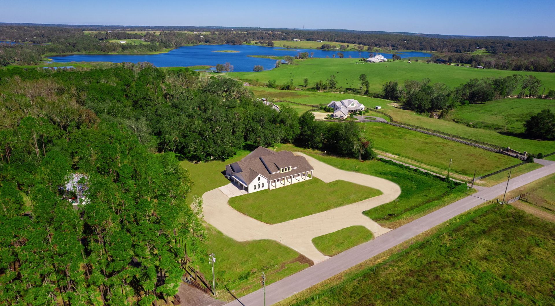 Aerial view of a house with a winding driveway, surrounded by green fields, trees, and a lake in the background.