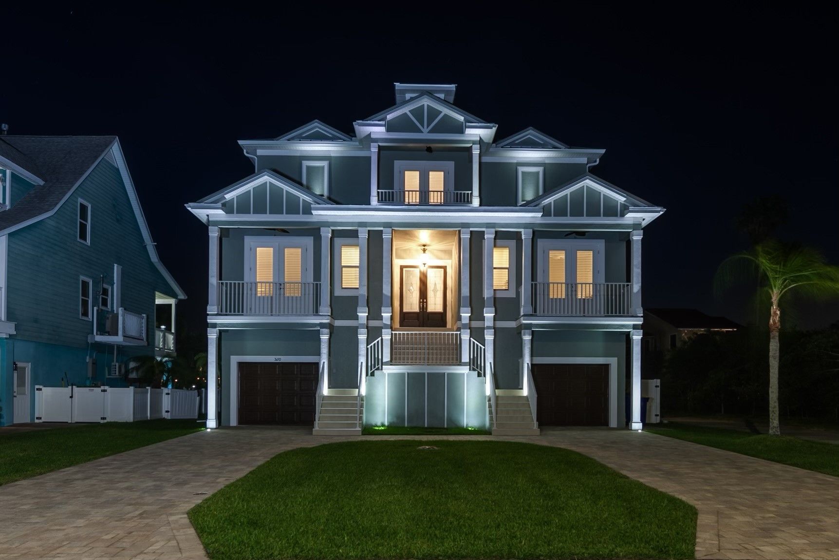 Three-story teal house at night, lit with white lights. Two-car garage, front lawn, and driveway.