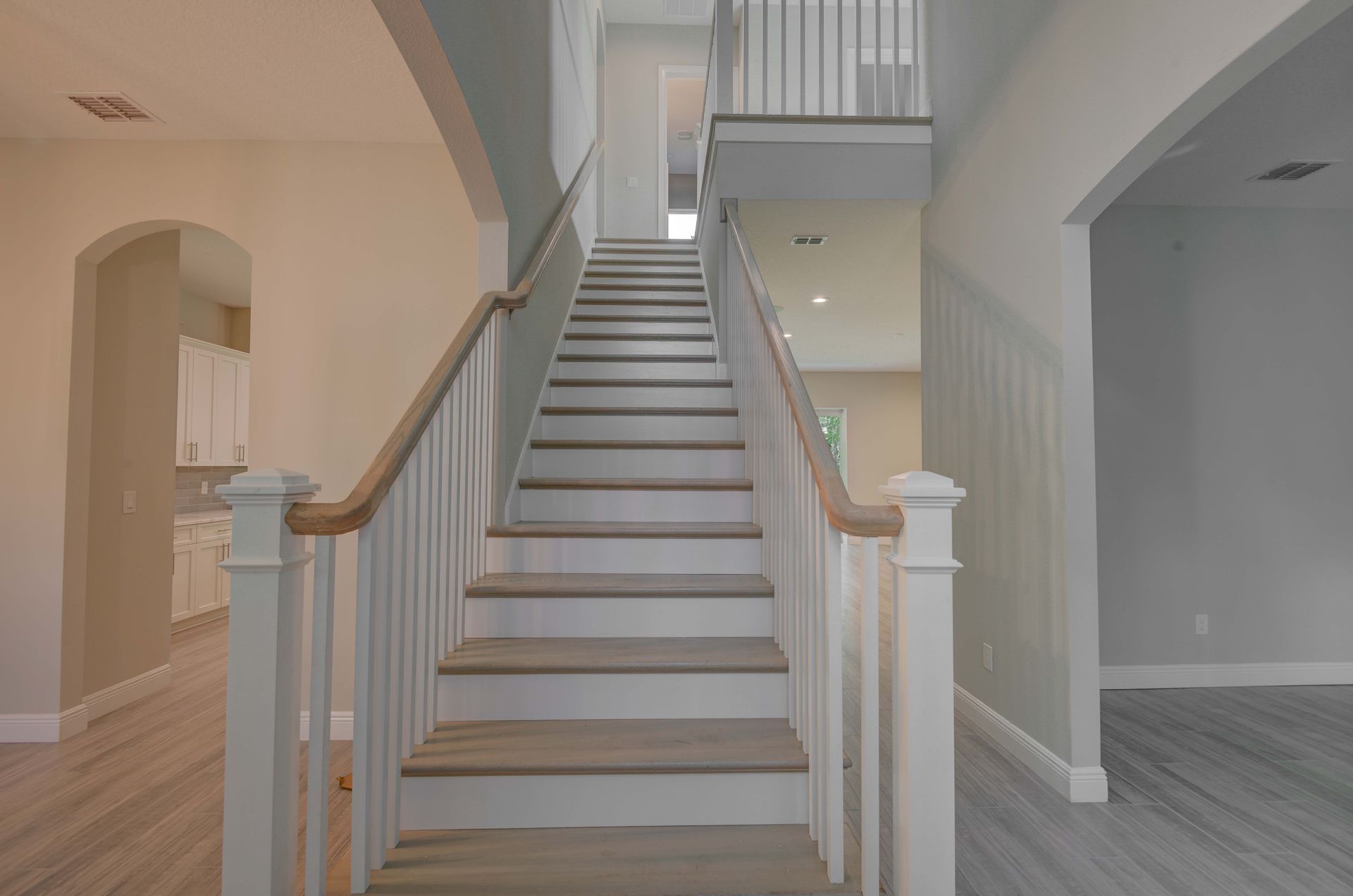 Staircase in a home with wooden handrails and light-colored walls. A kitchen and living area are visible.