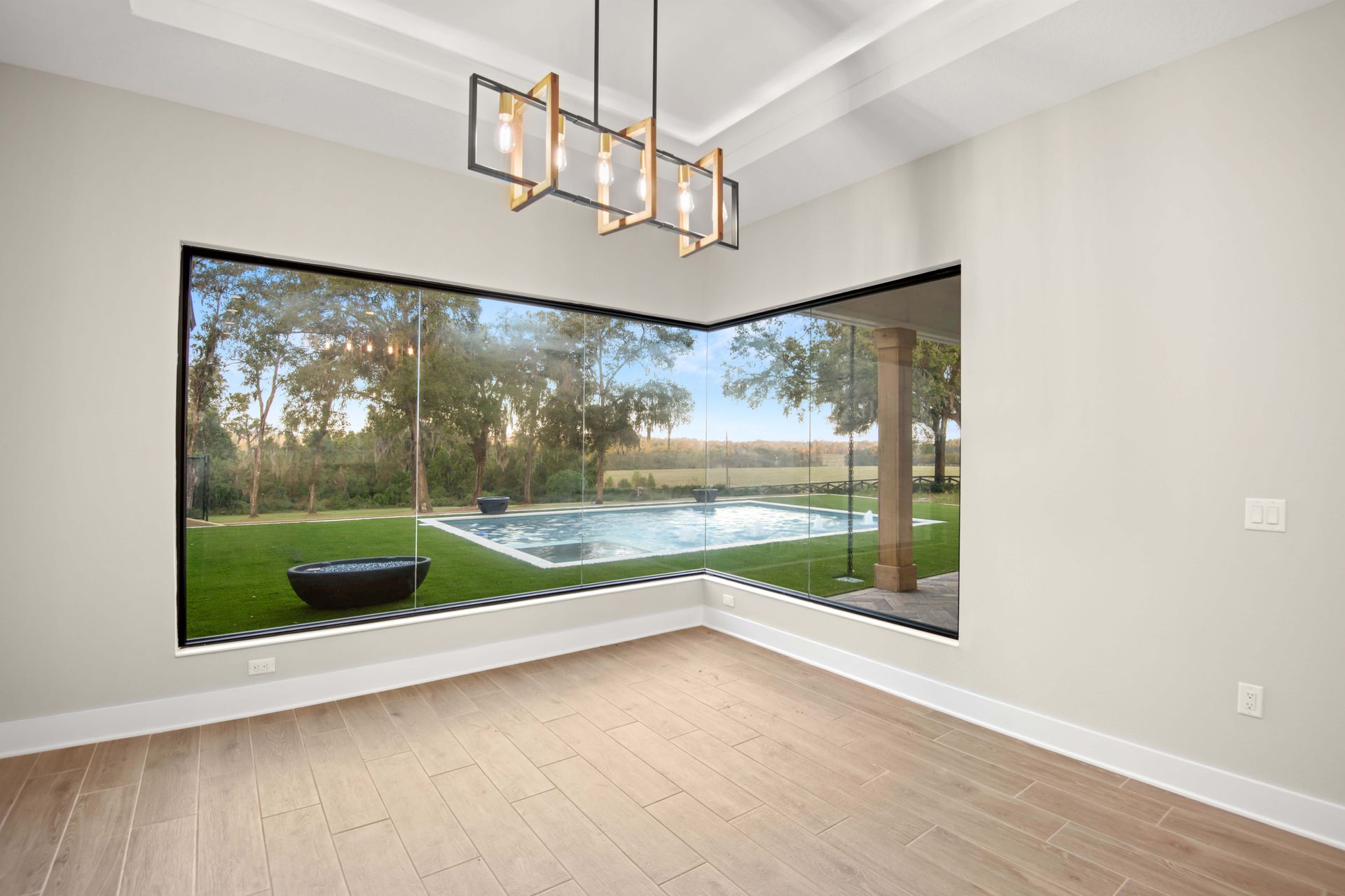 Empty room with large corner window overlooking a pool and yard, neutral walls, wood floor, and a modern chandelier.