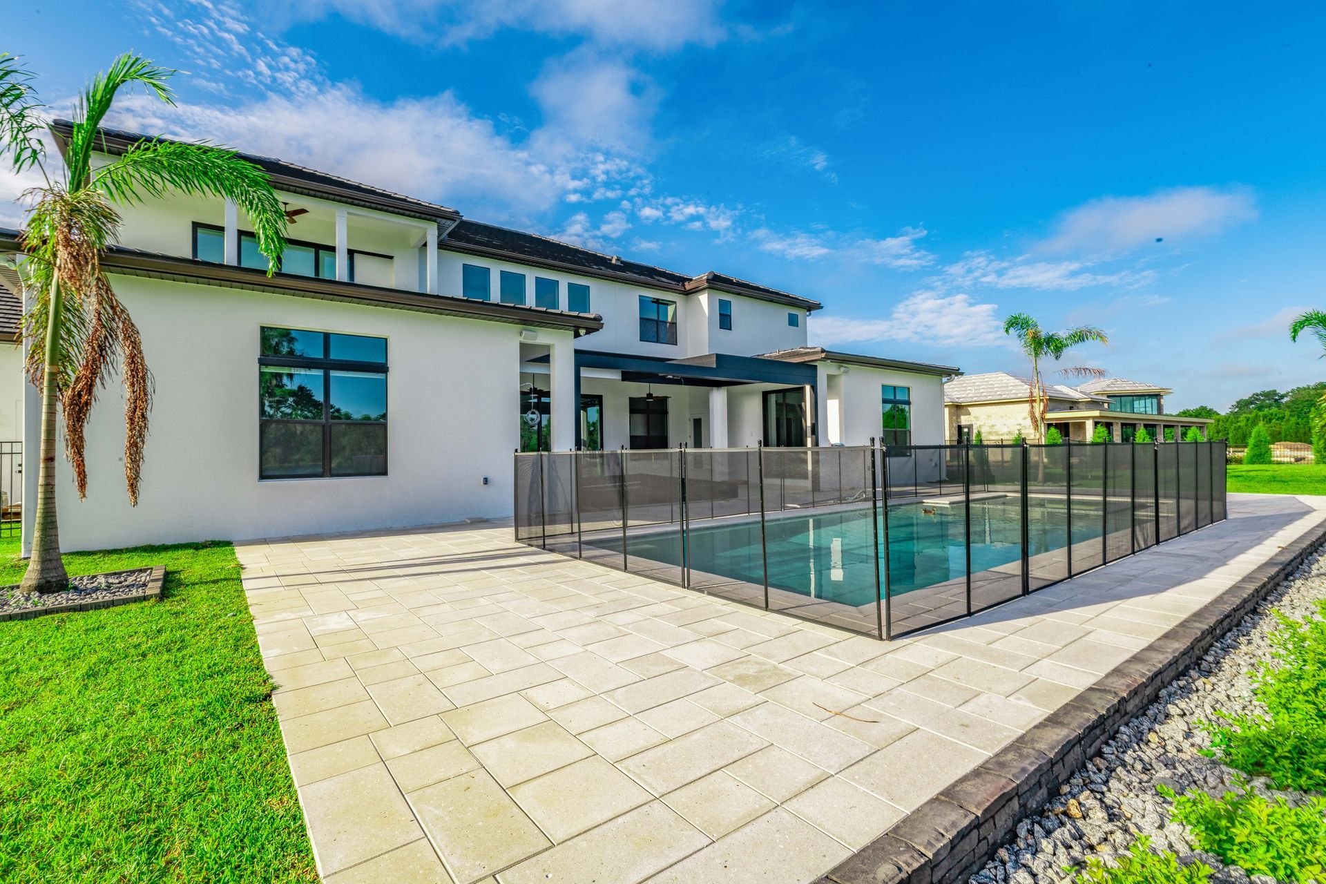 Backyard of a modern white house with a pool, safety fence, and patio on a sunny day.