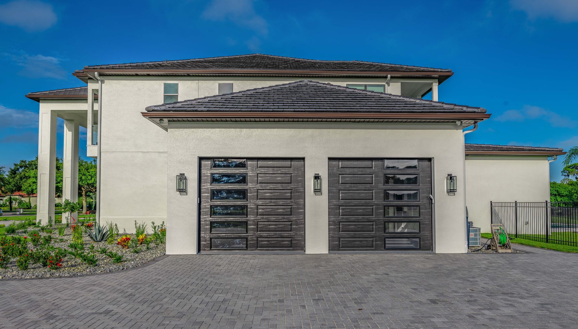 Two-story house with gray garage doors. Light-colored stucco exterior, dark roof, and blue sky.