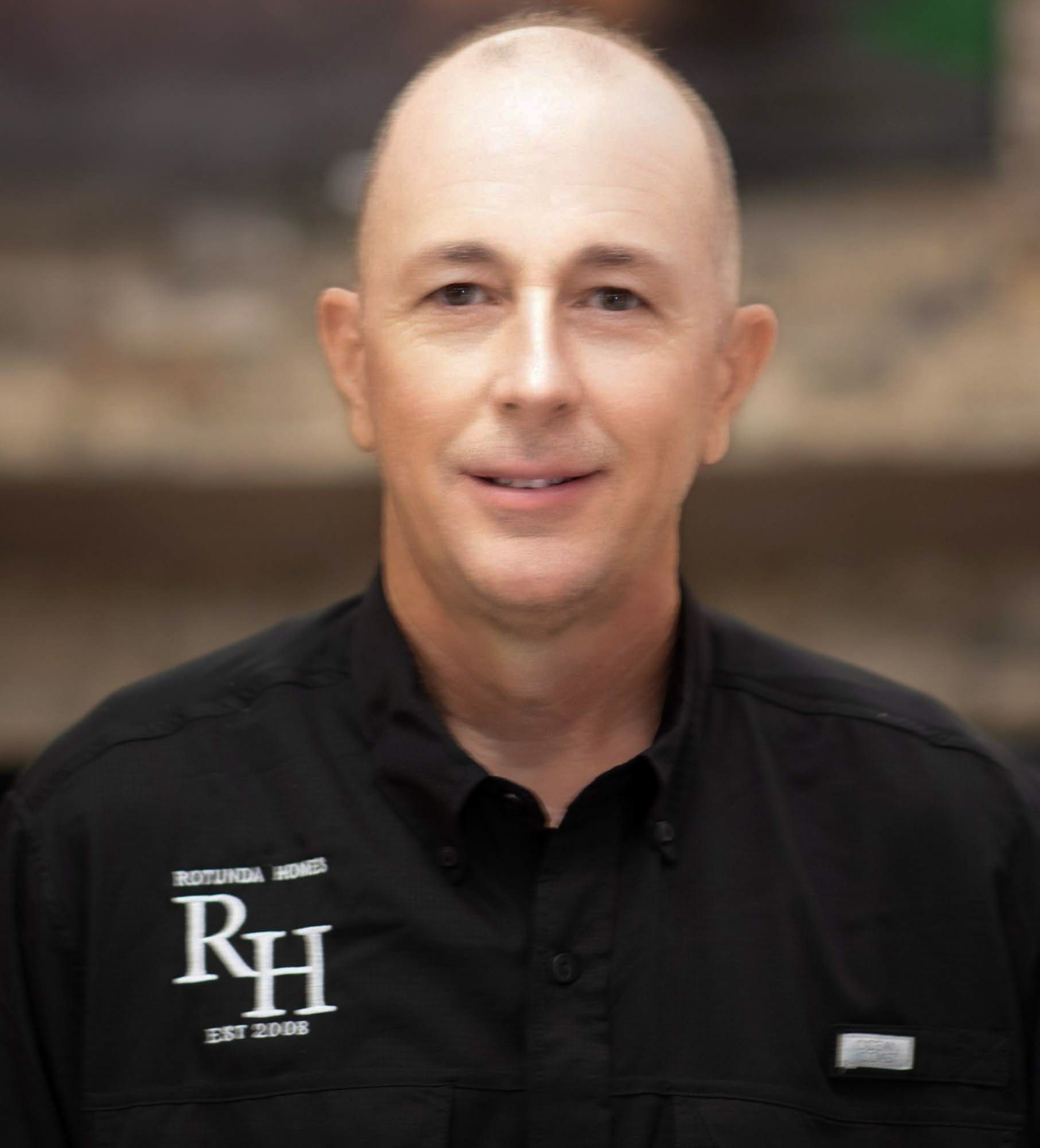 Bald man in black shirt with logo; smiling, indoors.