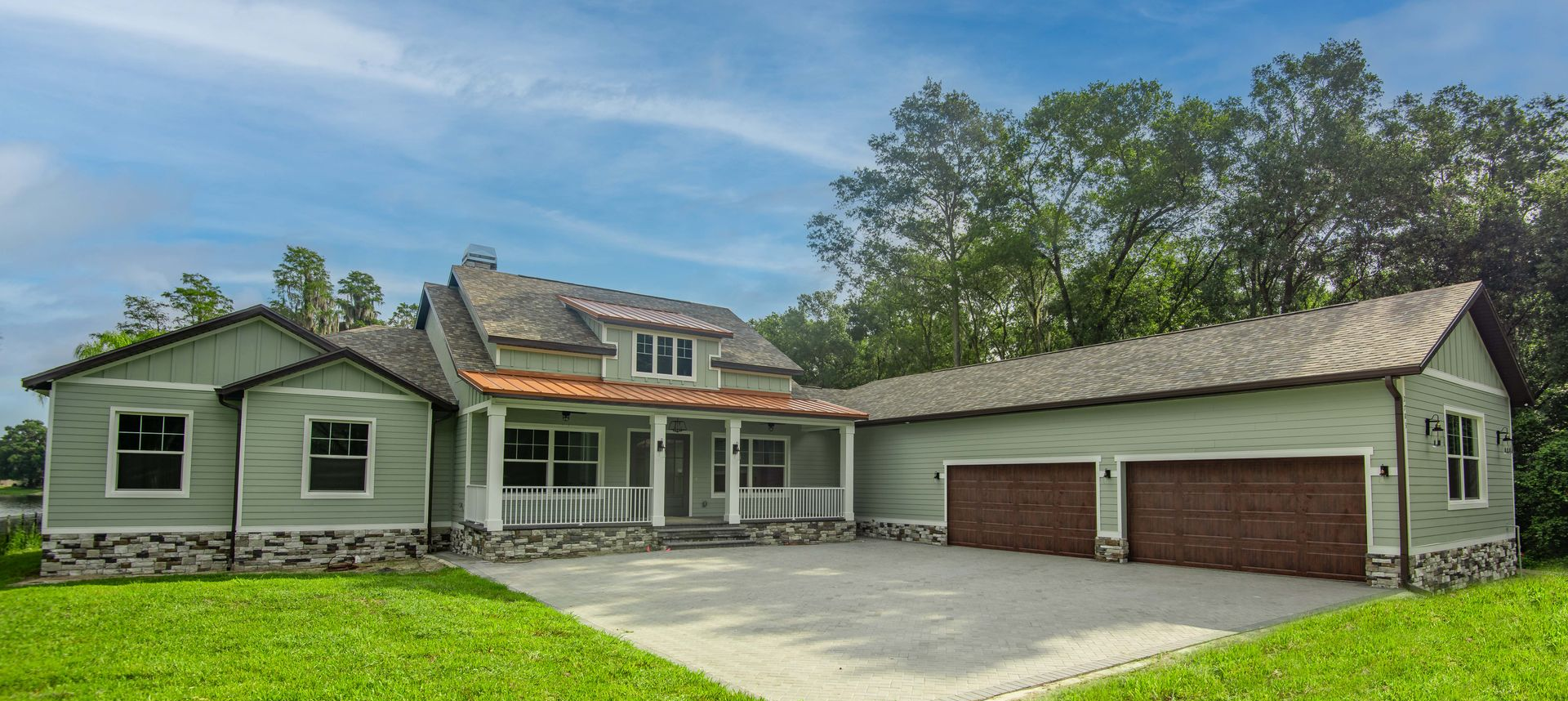A light green house with a gray roof and brick garage doors on a green lawn with trees.