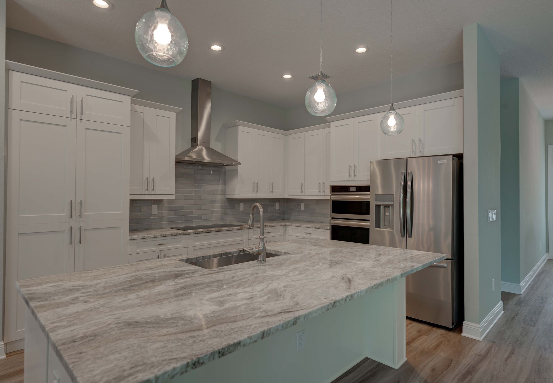 A modern kitchen with white cabinets, a large gray marbled island countertop, stainless steel appliances, and pendant lights.
