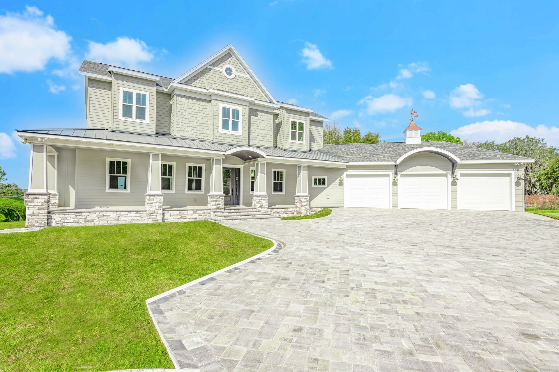 Two-story gray house with stone accents, a three-car garage, and a cobblestone driveway. Green lawn and blue sky.