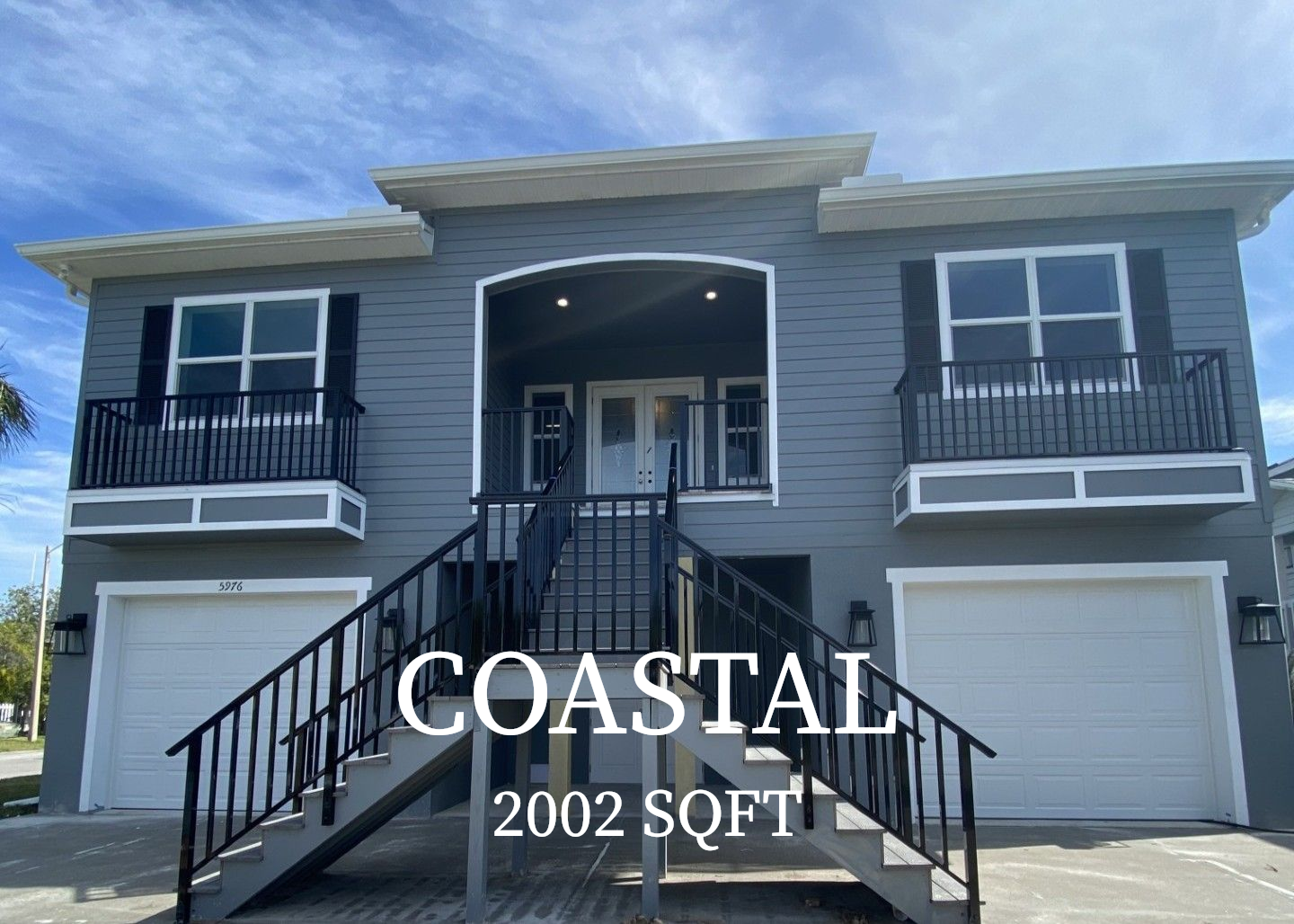 A gray two-story Coastal-style home with white trim, two garage doors, and a central staircase leading to a front entry.