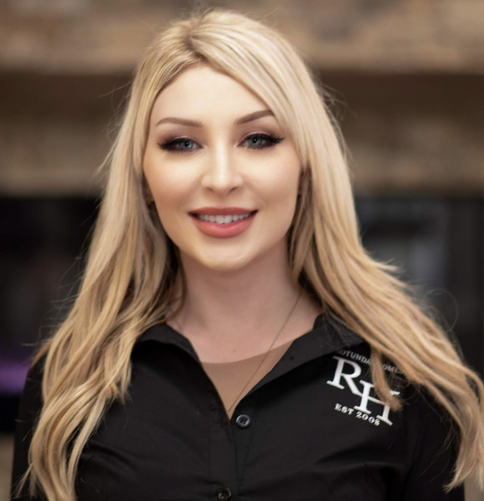 Blonde woman with dark eye makeup smiles, wearing a black shirt with logo.