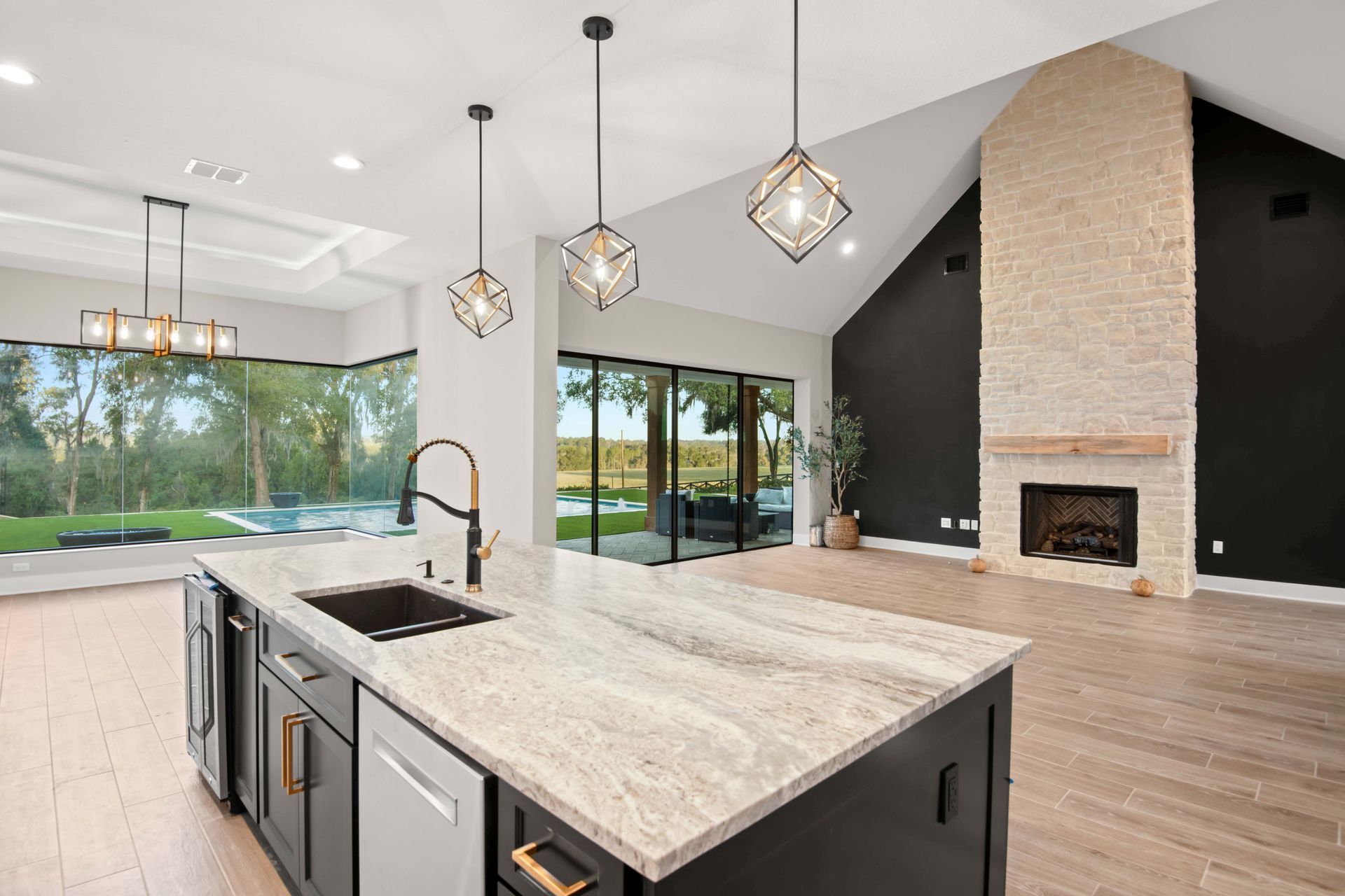 Spacious kitchen with island, pendant lights, large windows overlooking a backyard and a fireplace on a black wall.