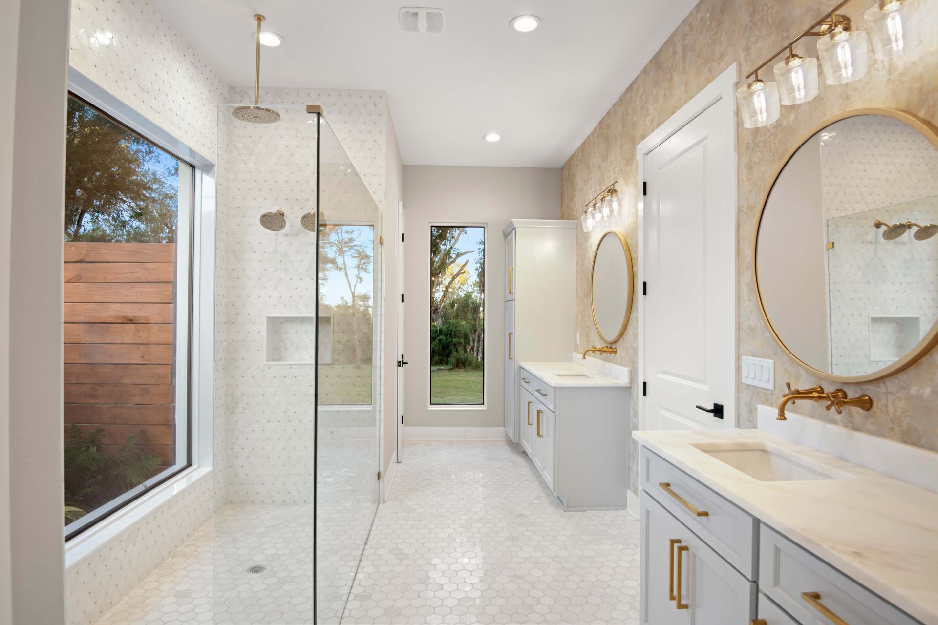 Bright, modern bathroom with glass shower, double vanity, and large windows. White and gold accents.