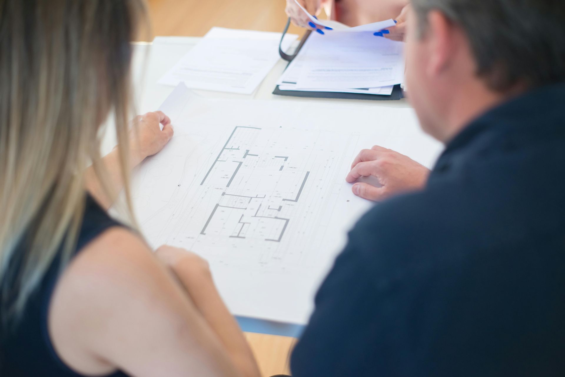 People looking at blueprints on a white table. Light-colored hair, dark shirt, papers held.