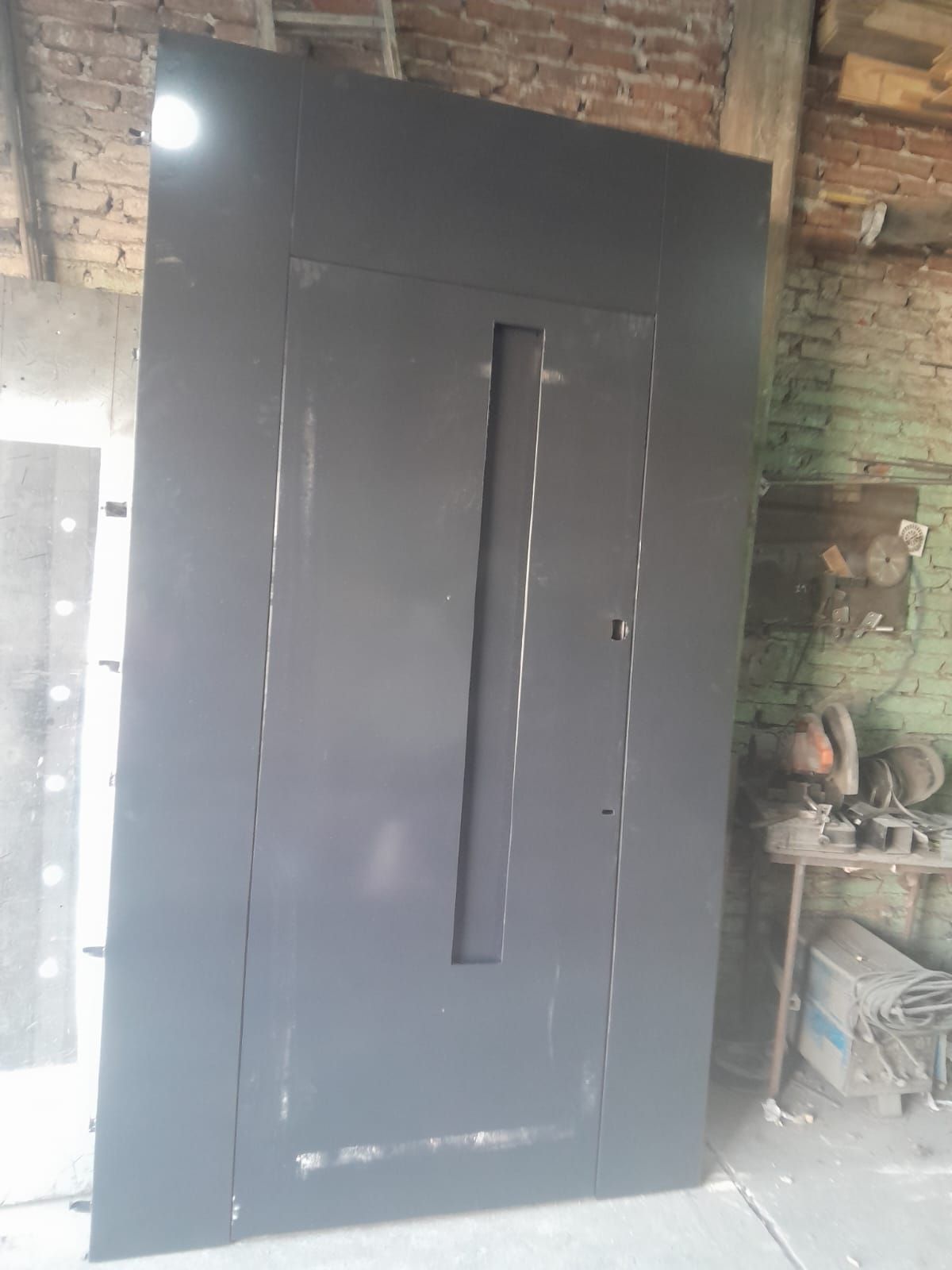 Gran puerta o portón de metal gris oscuro con una estrecha ventana vertical en un entorno de taller rústico.