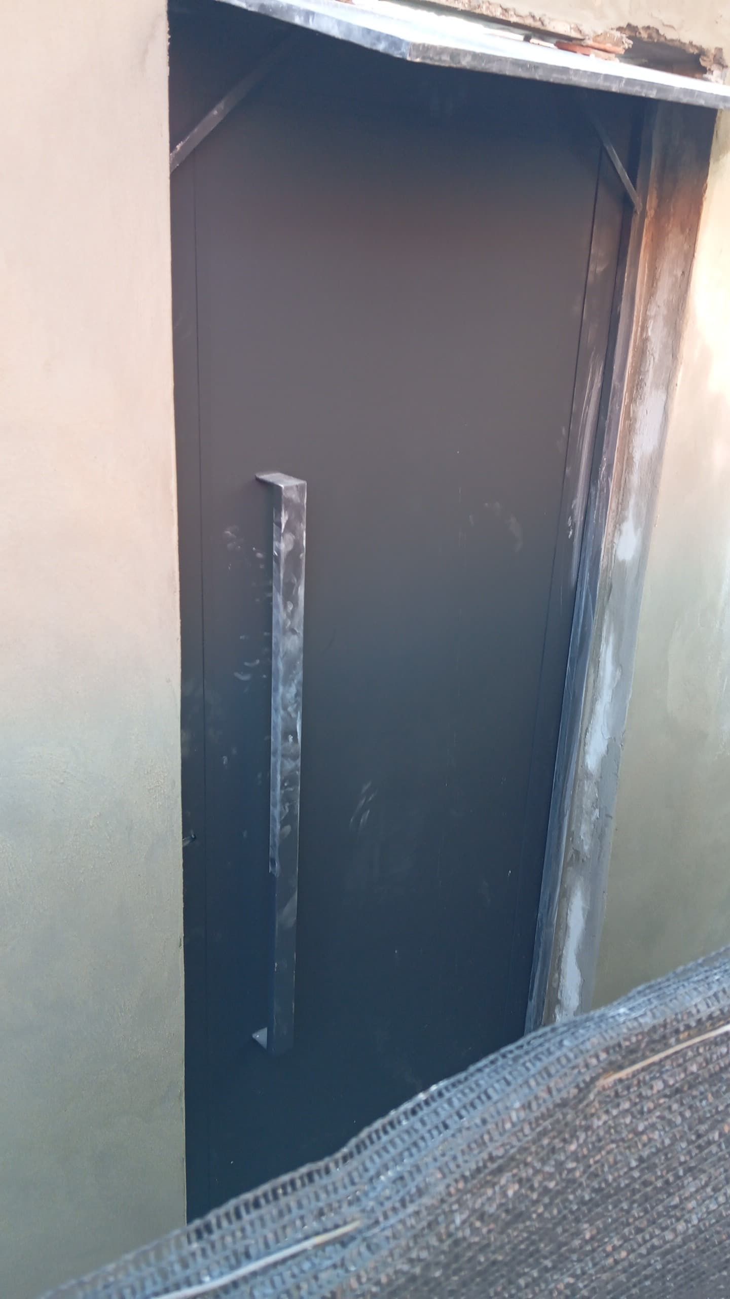 Una estrecha puerta o trampilla oscura en una pared de color beige, vista desde el exterior a la luz del día.