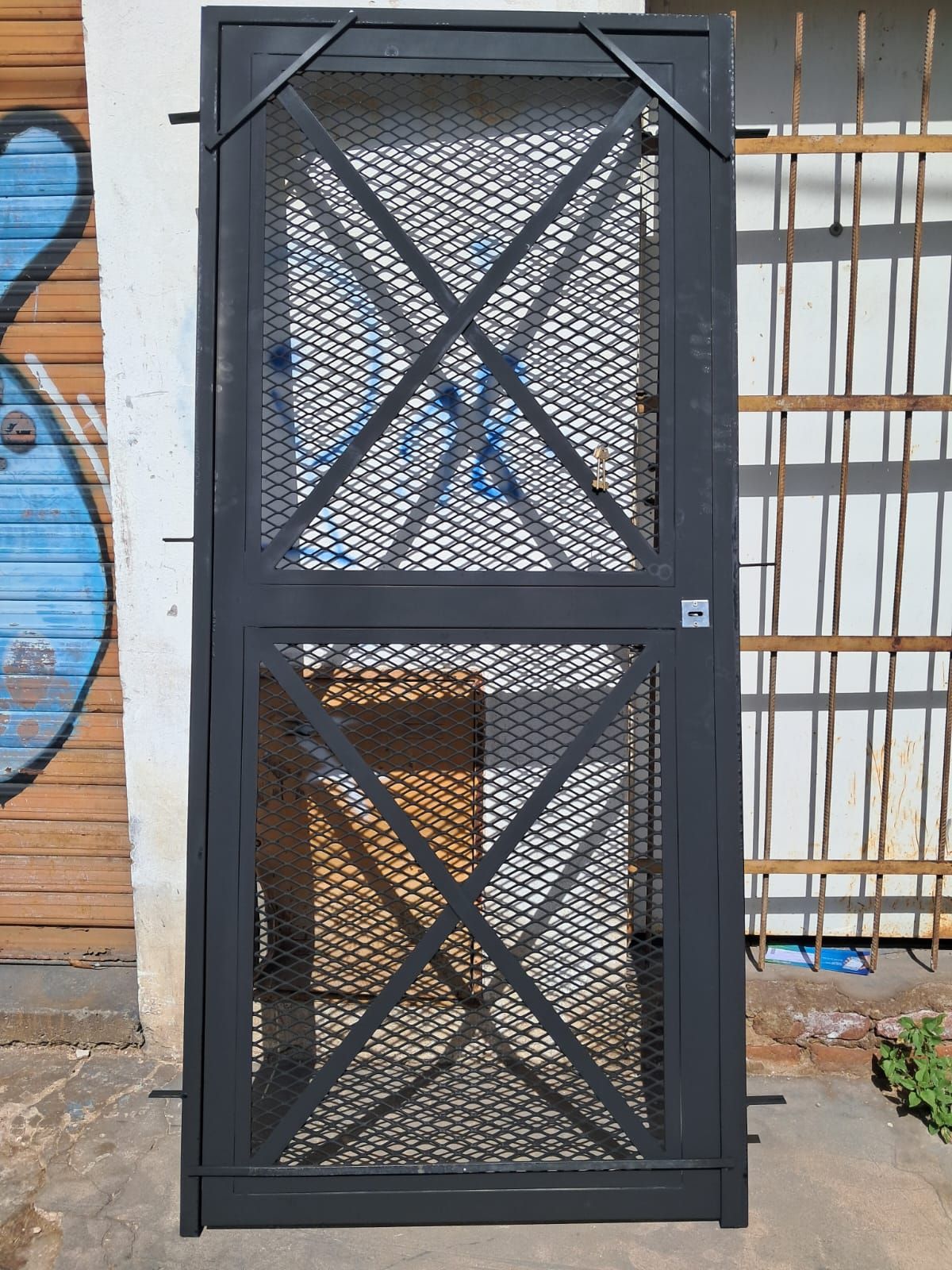 Puerta de seguridad de malla metálica negra con refuerzos diagonales en forma de X, montada en el exterior contra una pared.