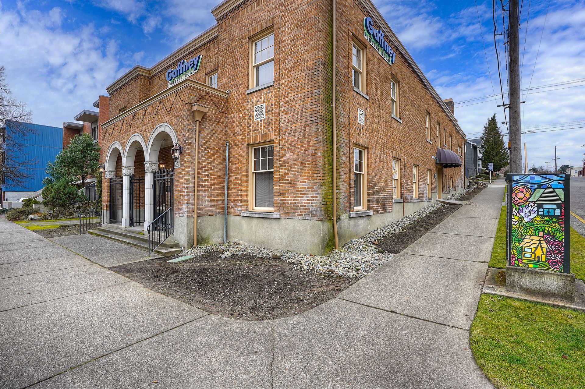 Brick building with arched doorways at a street corner, beside a sidewalk and utility pole under a blue sky