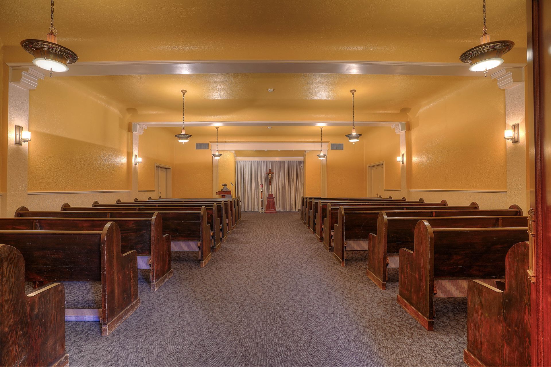 Empty chapel interior with wooden pews, carpeted aisle, and warm yellow lighting