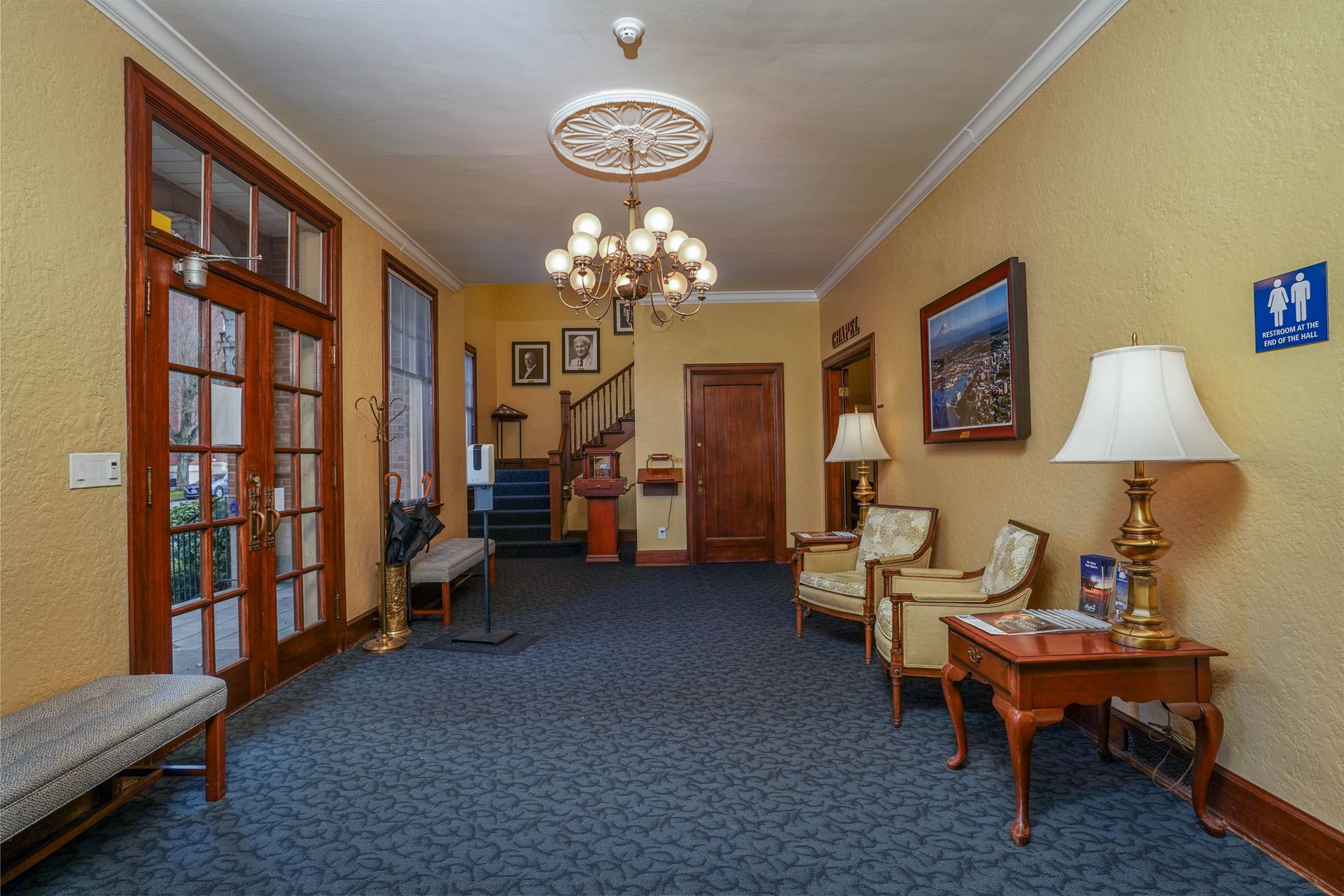 Elegant hallway with blue carpet, chandeliers, framed art, and wooden furniture along the walls