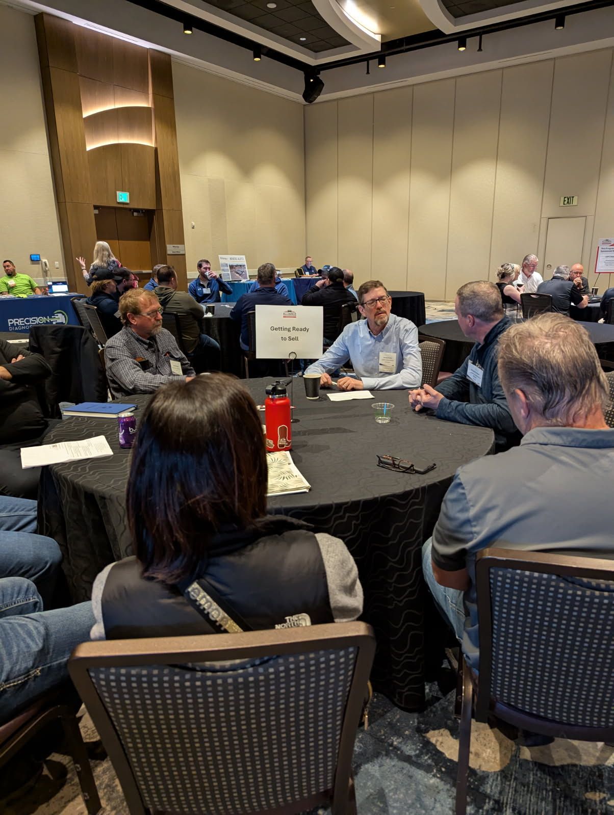 People seated at a round table, possibly in a conference or meeting. One man is speaking.