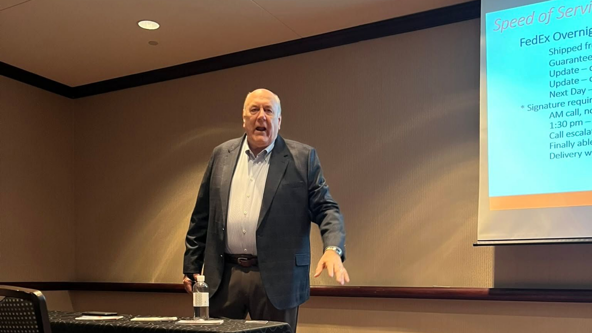 Man presenting on a stage beside a projected slide in a conference room
