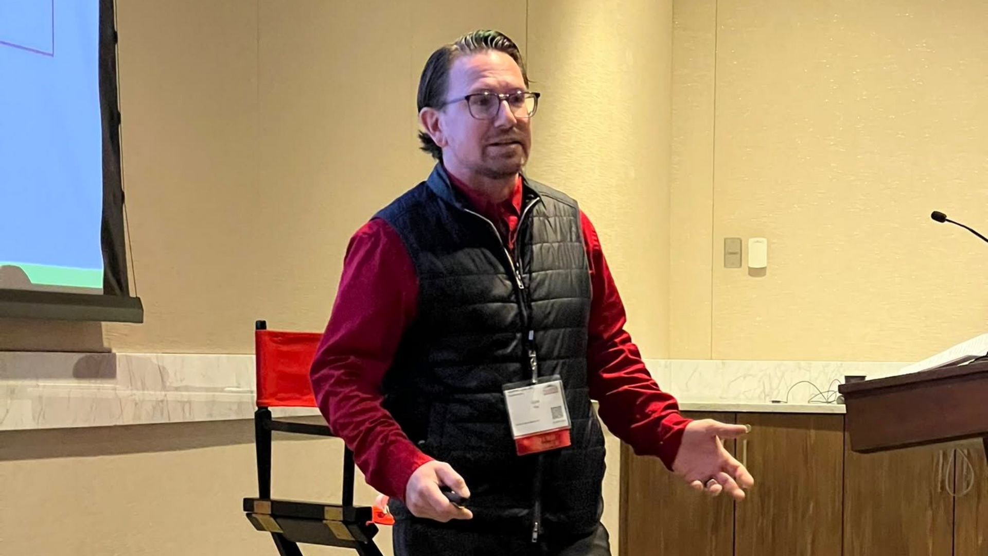 Speaker in a red shirt and black vest presenting in a lecture room, gesturing beside a podium.
