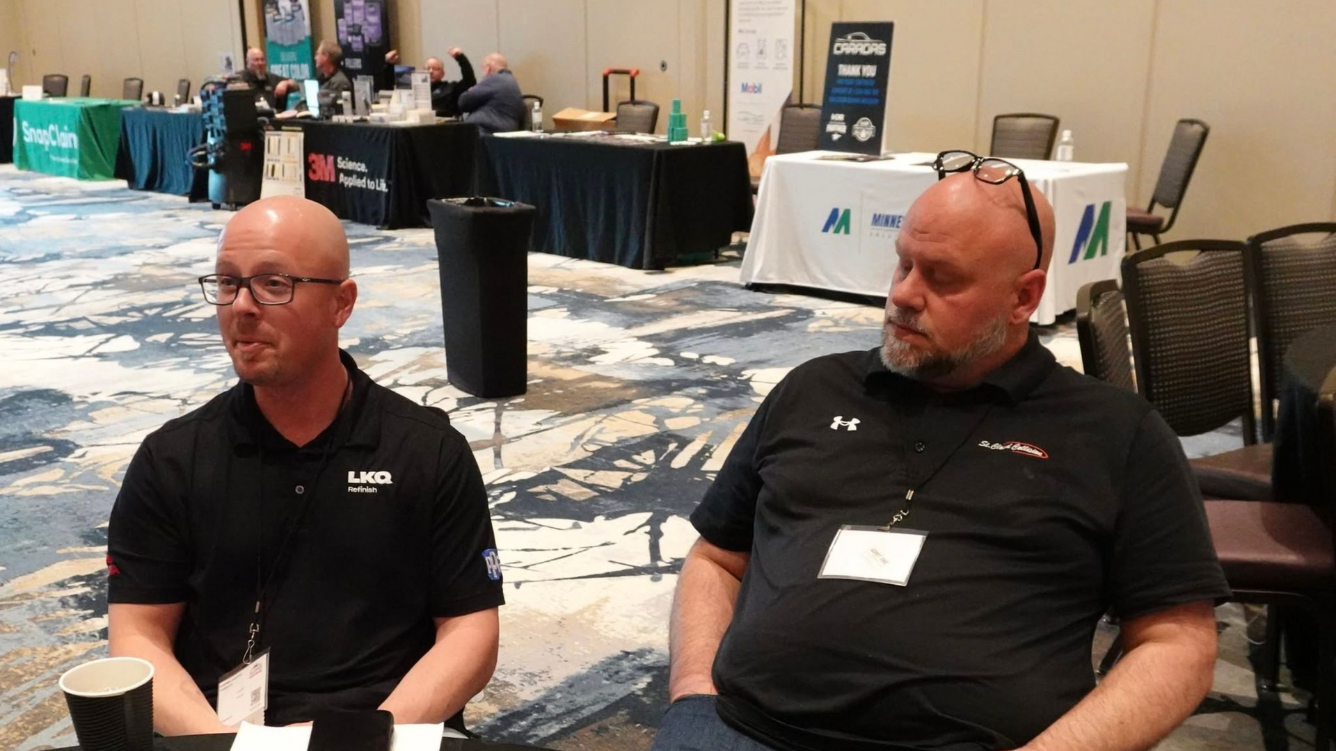 Two seated men in black shirts at a conference with tables and booths in the background