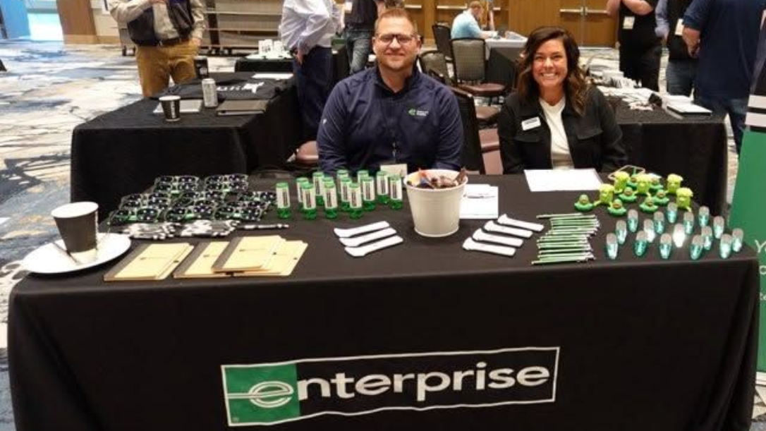 Enterprise booth with two smiling staff behind a table of giveaway items at a trade show